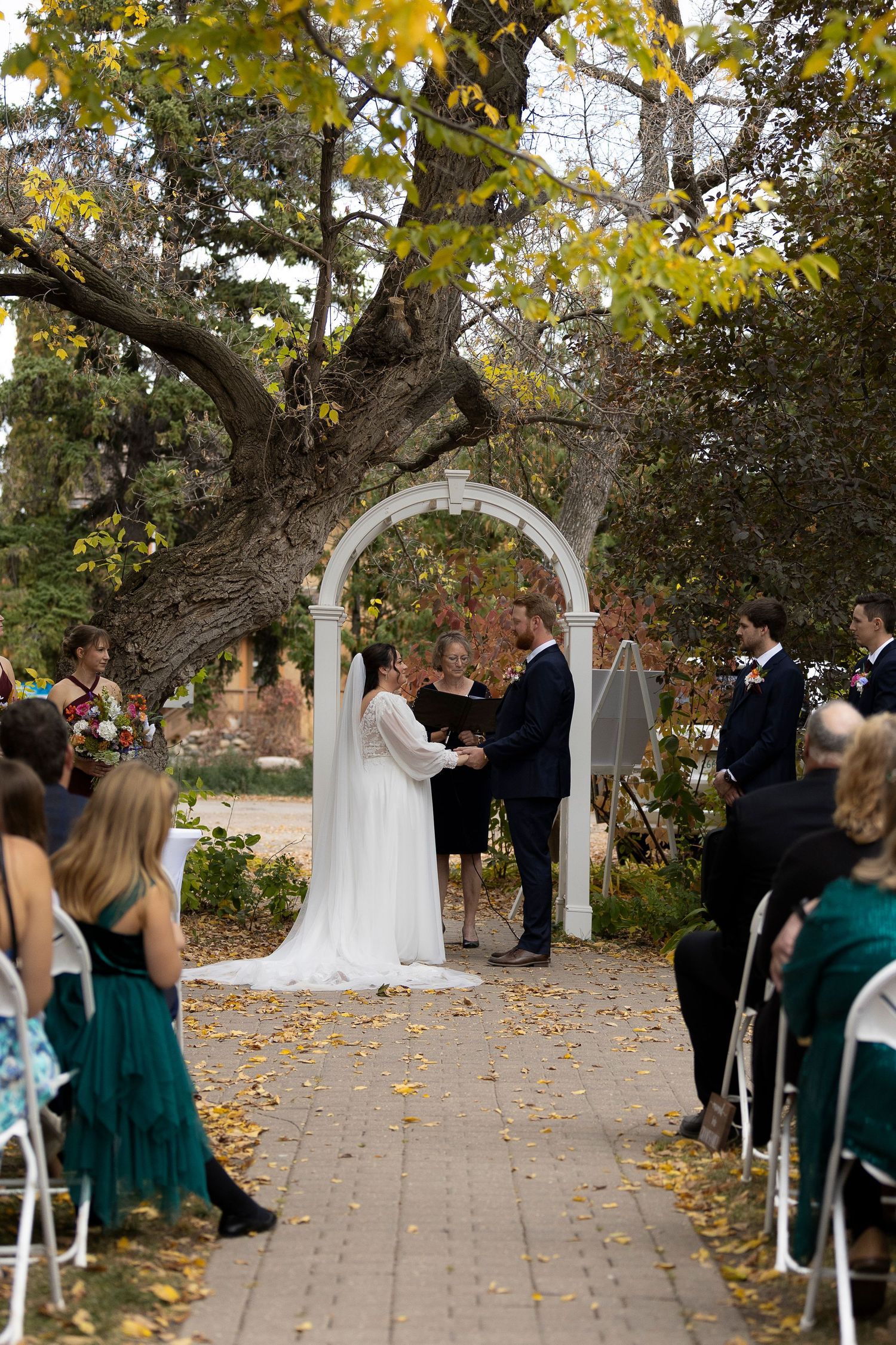 Outdoor wedding ceremony under white arch with fall foliage and guests seated at the Forestry Farm in Saskatoon, Saskatchewan.
