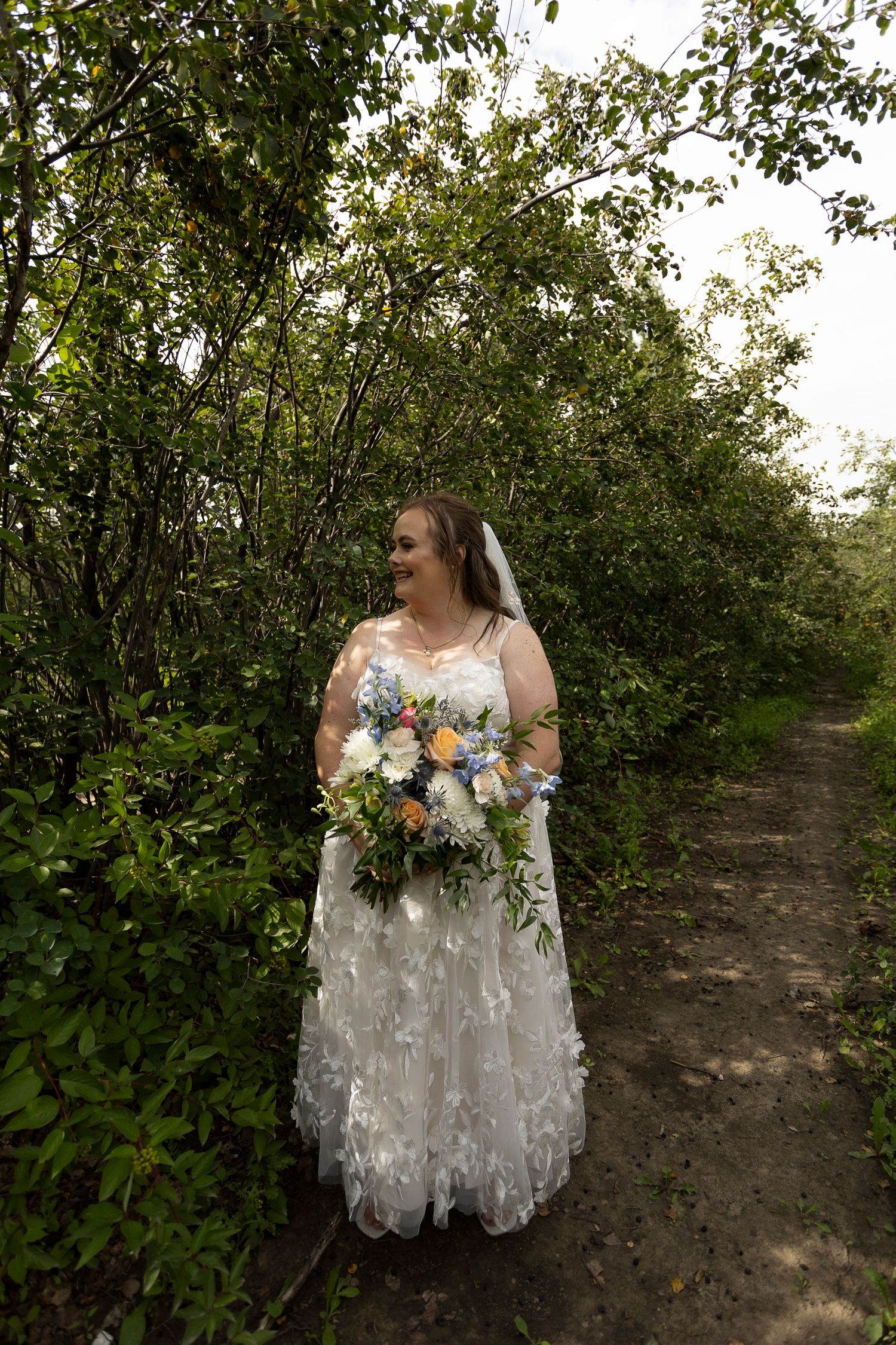 Bride in lace wedding dress holding colorful bouquet standing on garden path surrounded by lush green trees.