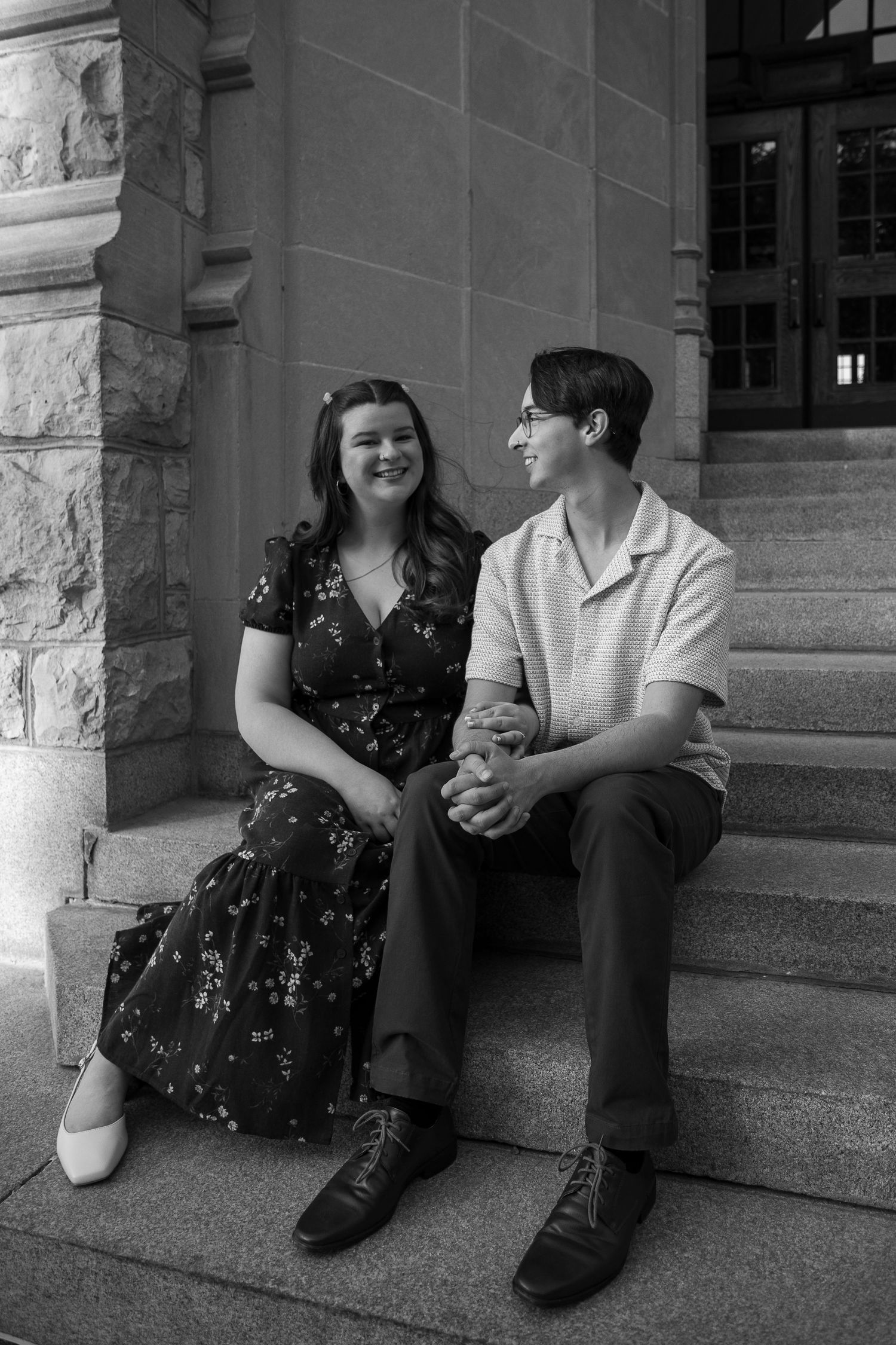 Black and white photo of a happy couple holding hands on stone steps outside a classic stone building.