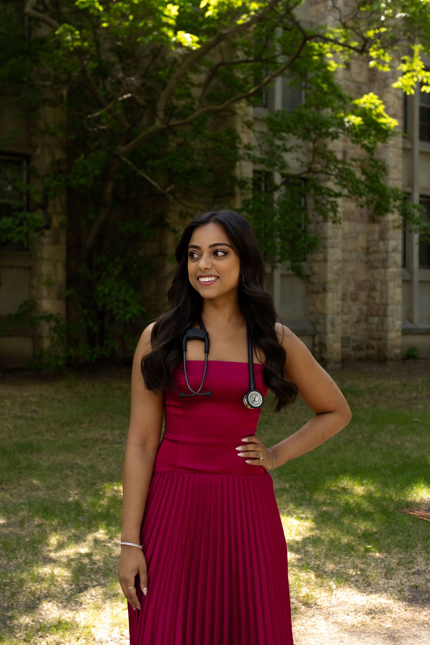 Young woman in red pleated dress with stethoscope smiling outdoors near stone building.