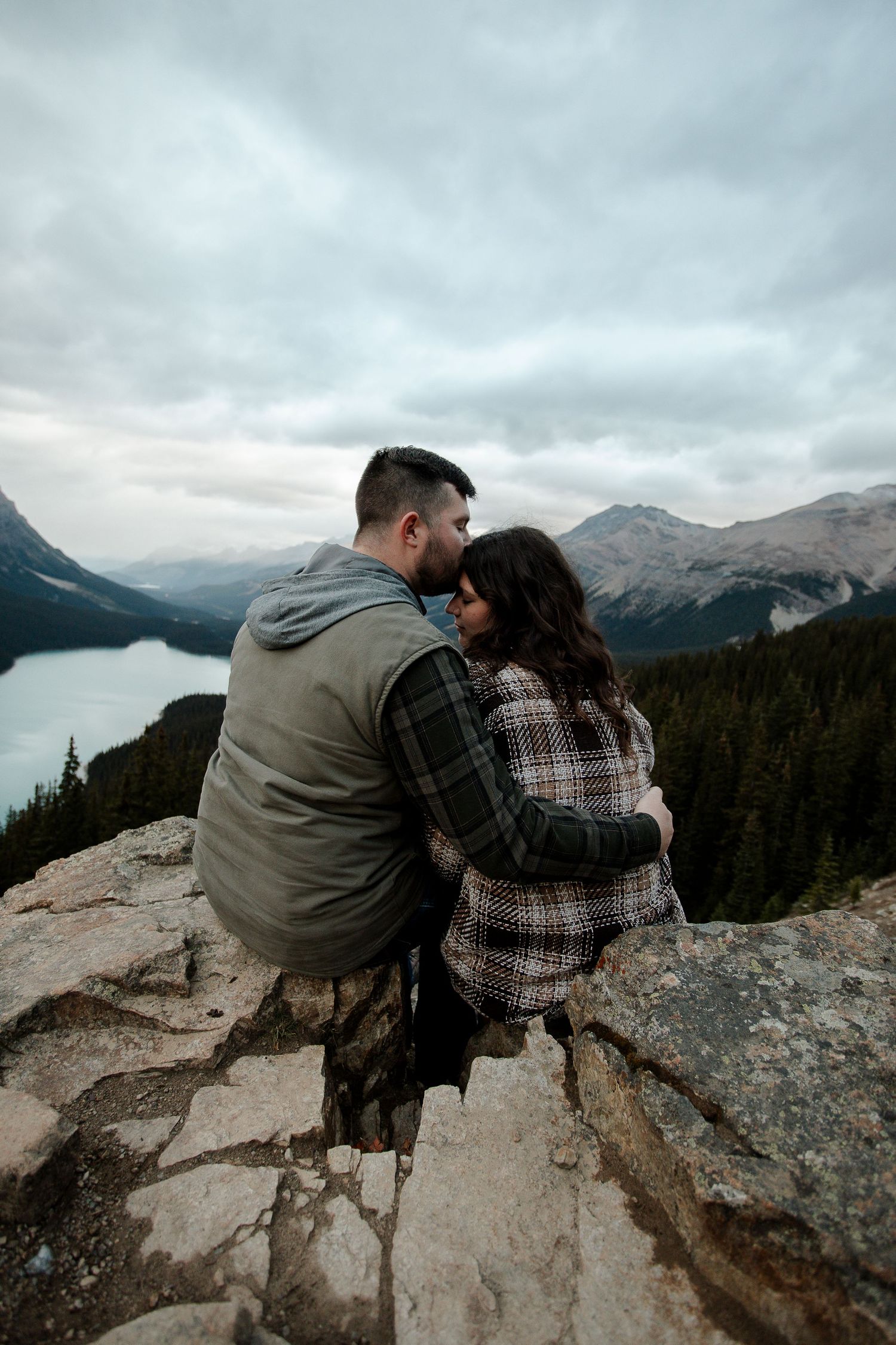 Couple embracing on rocky mountain overlook with turquoise lake and misty peaks in background.