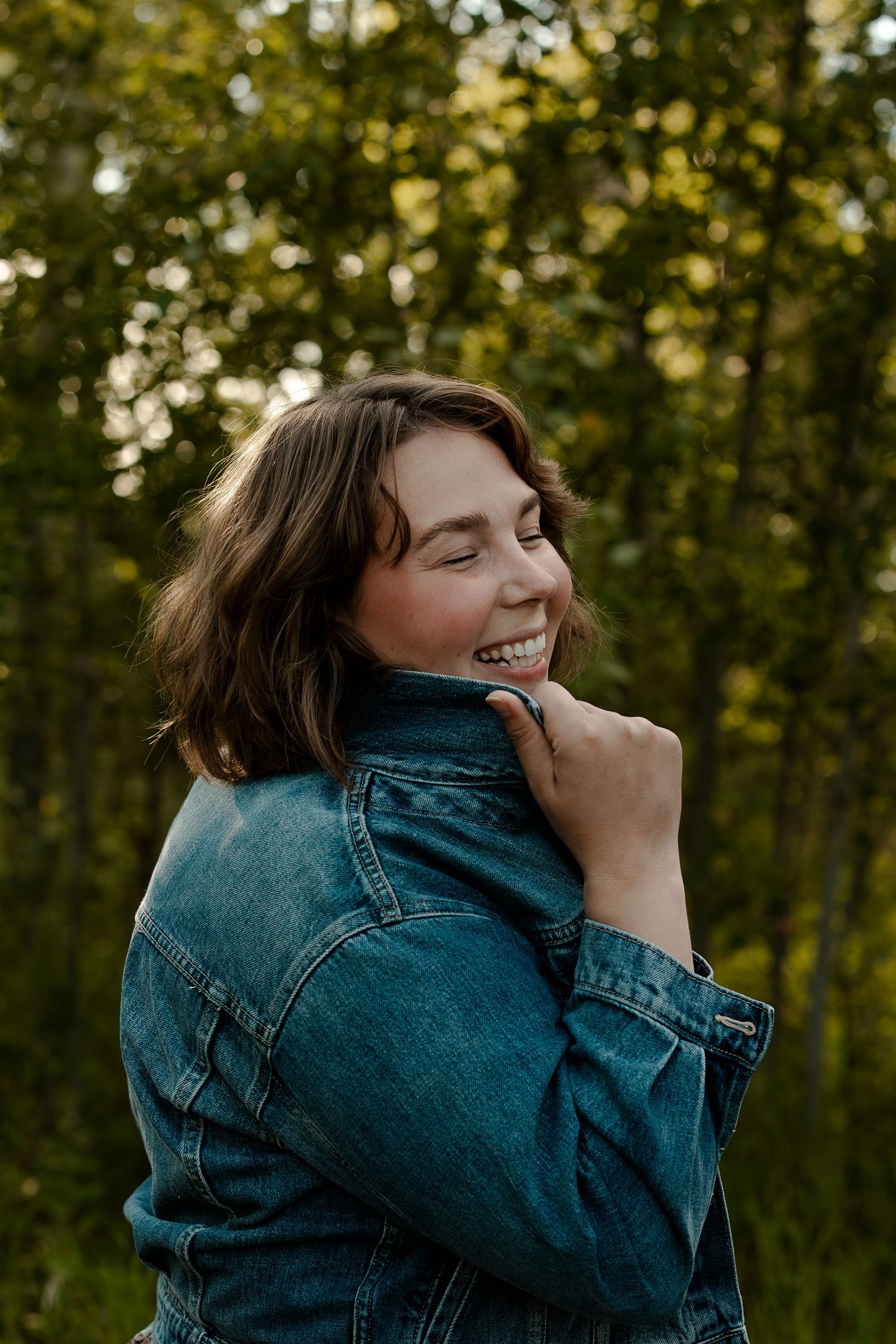 Casual outdoor portrait with denim jacket against green foliage at sunset.