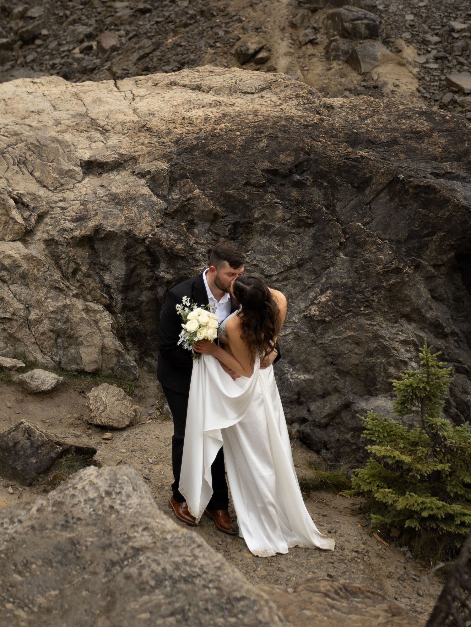 Bride and groom share a romantic kiss in wedding attire against a rugged rocky mountain backdrop.