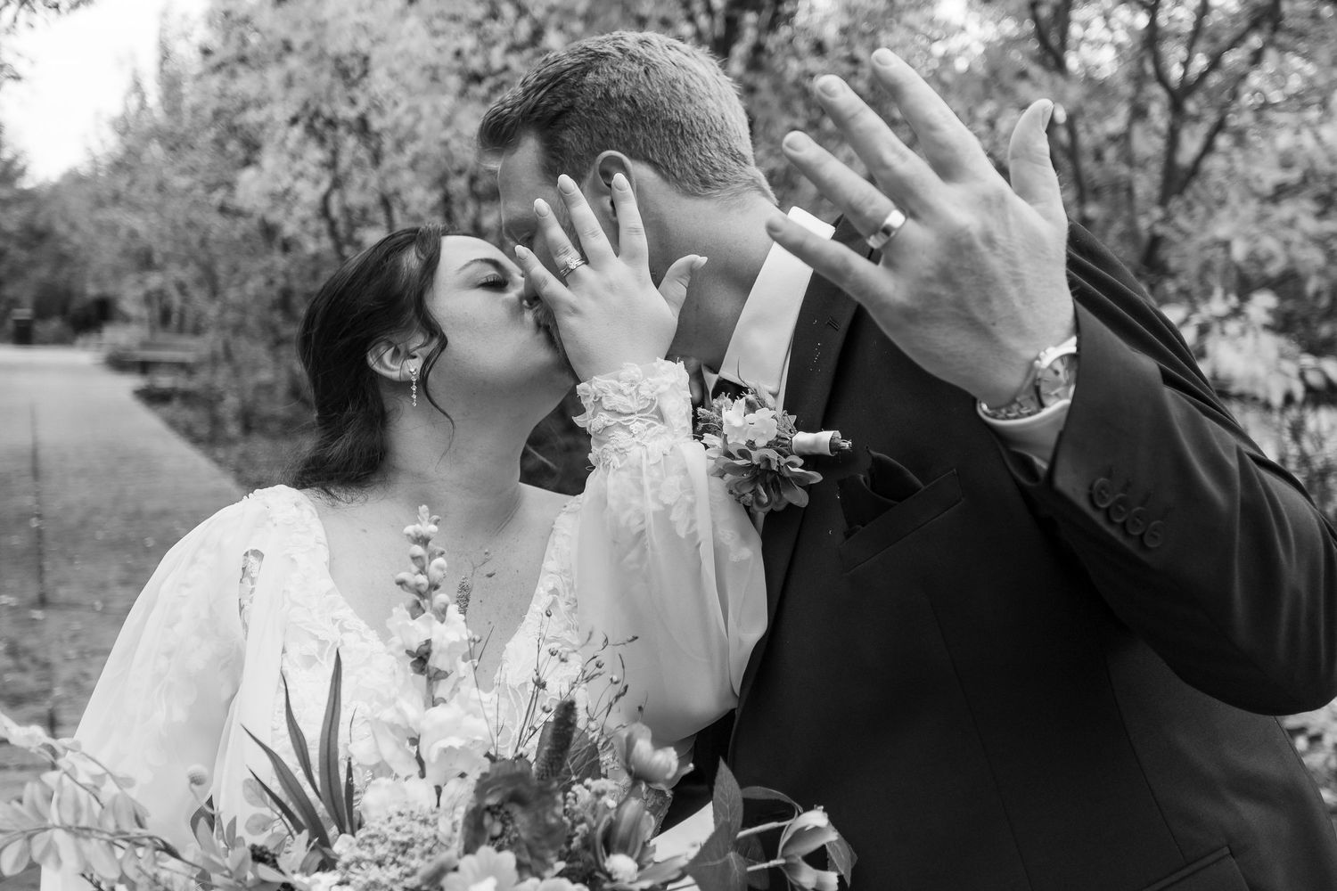 Black and white photo of newlyweds kissing while showing off wedding rings, bride holding bouquet outdoors.