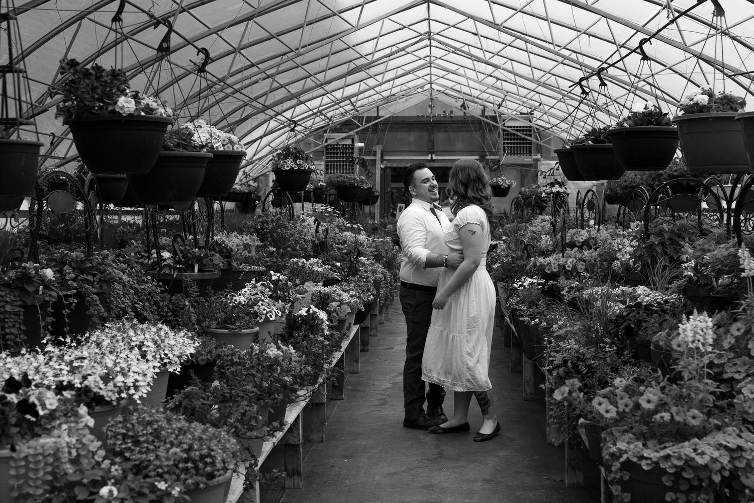 Black and white photo of a couple embracing in a lush greenhouse filled with hanging plants and flowers.