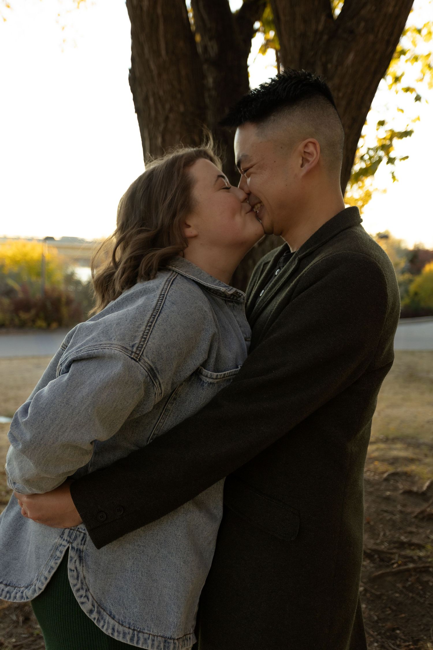 Couple sharing a romantic moment, nearly kissing outdoors near a large tree during golden hour.