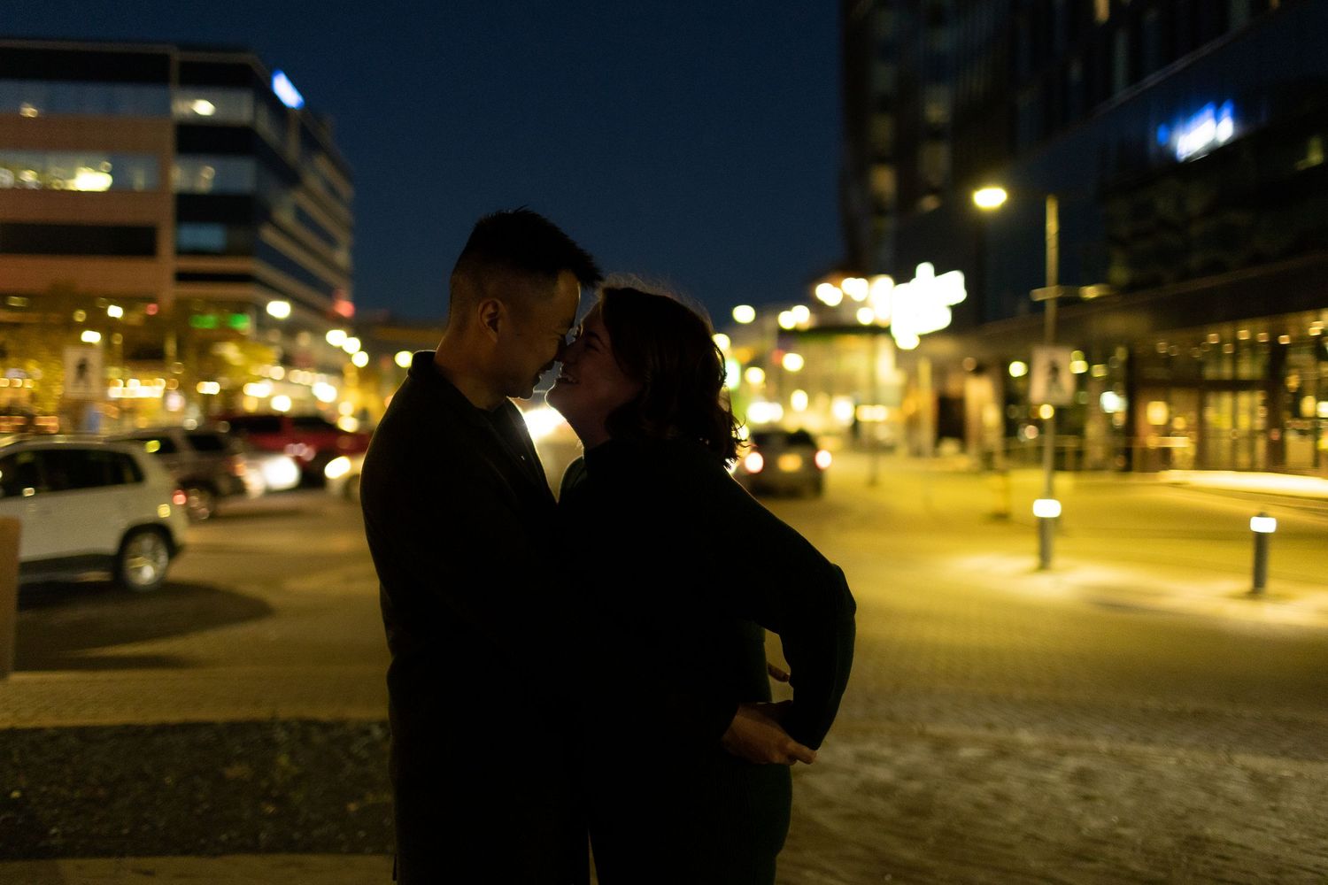 Romantic couple sharing a kiss at night in an illuminated urban plaza with city lights glowing behind them.