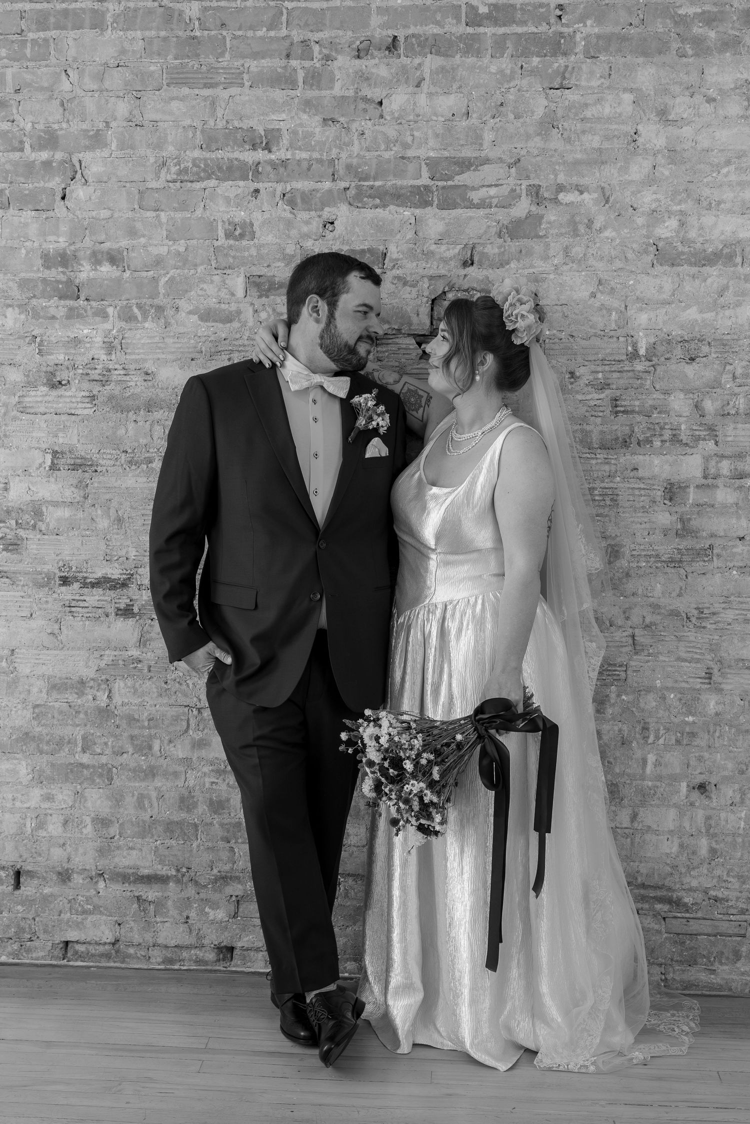 Black and white wedding photo of bride and groom smiling at each other against a rustic brick wall.