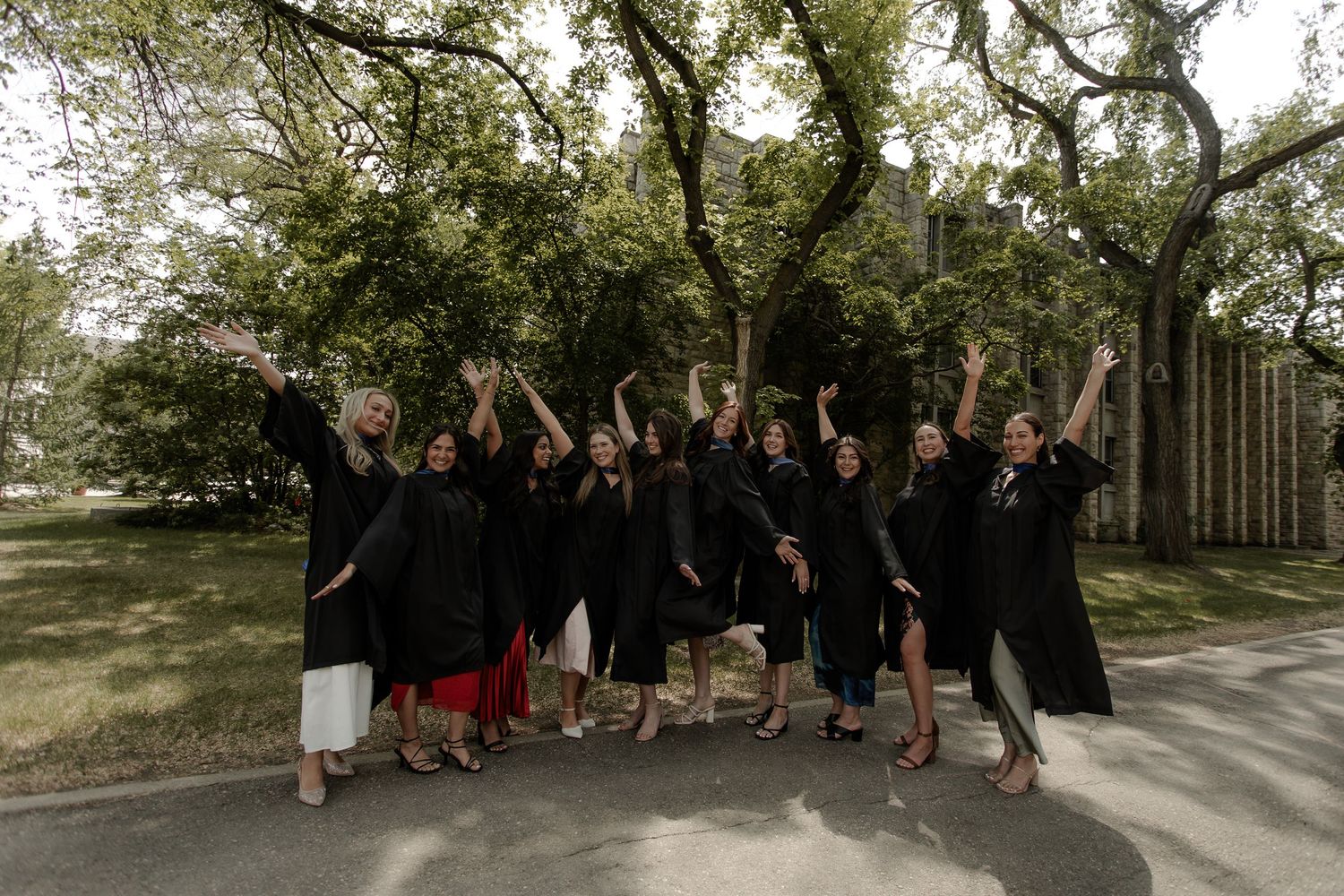 Excited graduates in black gowns cheering together outdoors on a sunny day near trees.