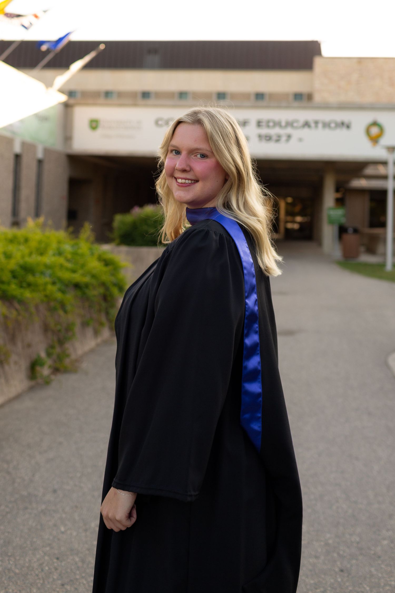 Smiling female graduate in black gown and blue sash poses outside College of Education building on graduation day.