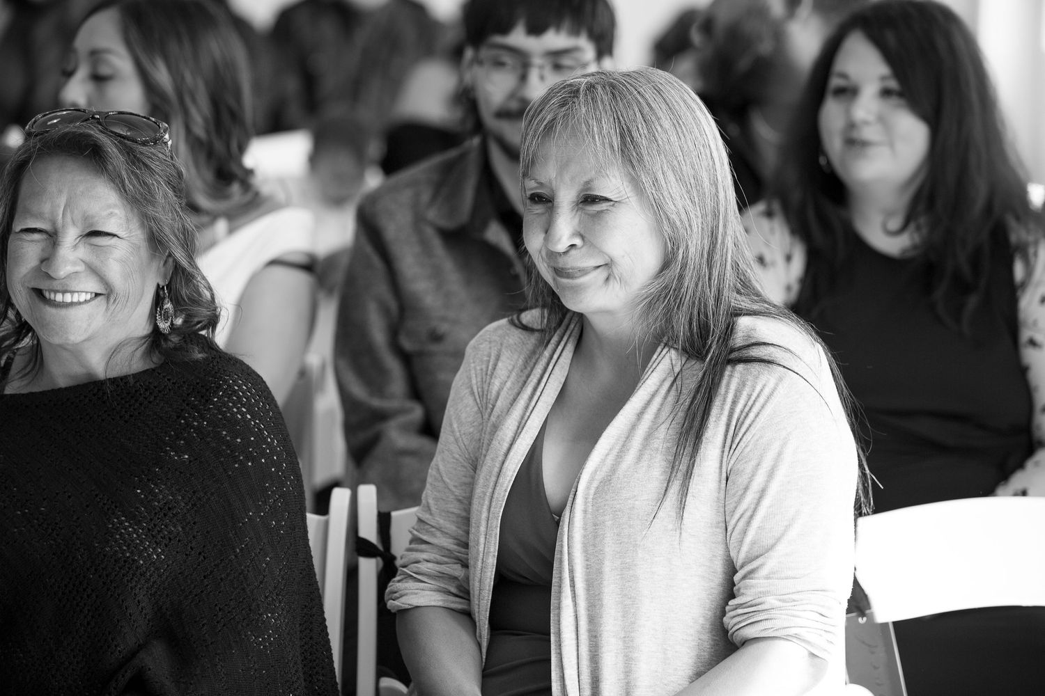 Black and white photo of smiling women sitting at an indoor event, enjoying a casual gathering.