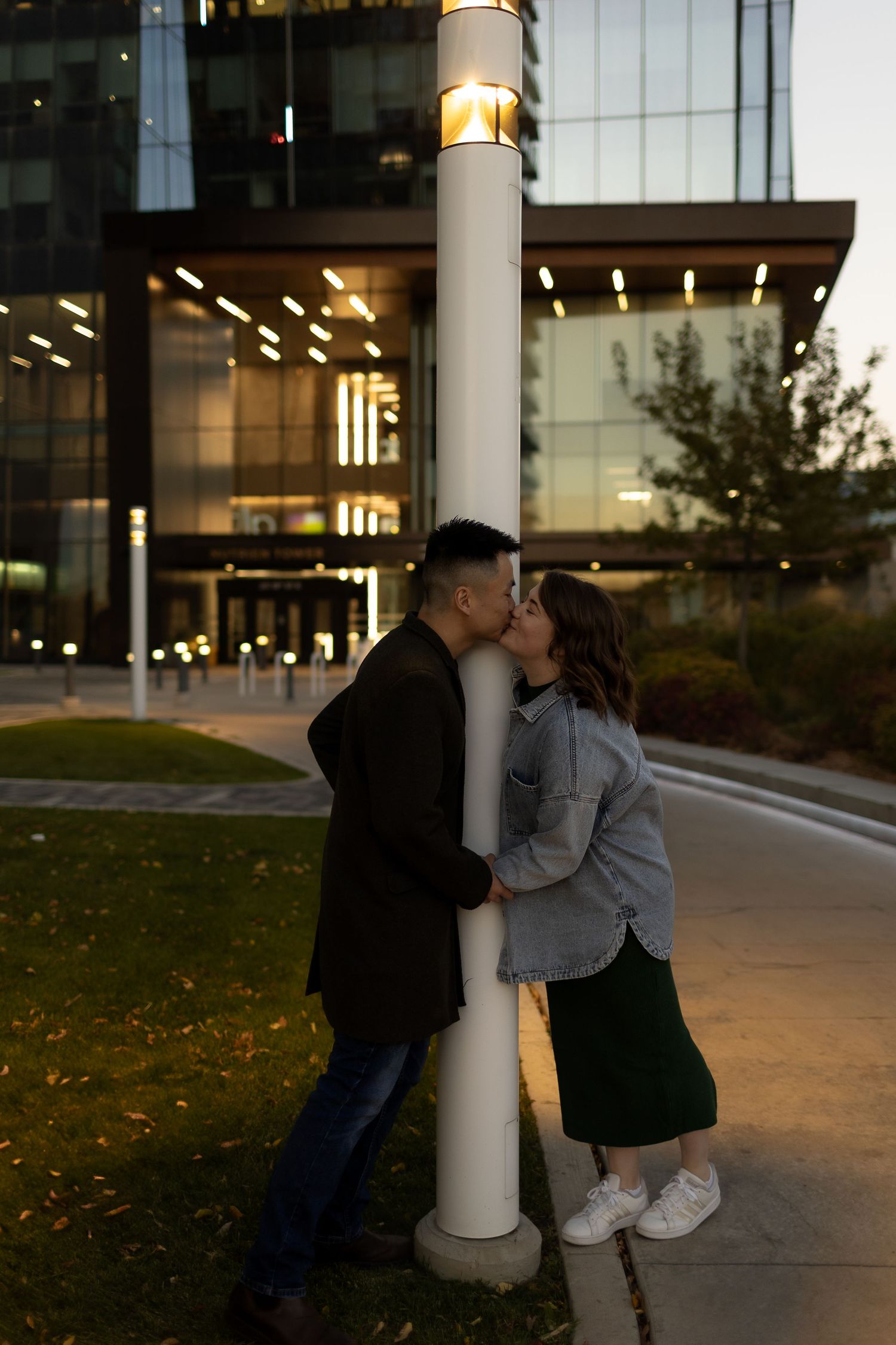 Couple sharing a romantic kiss behind a glowing lamppost outside a modern glass building at dusk.