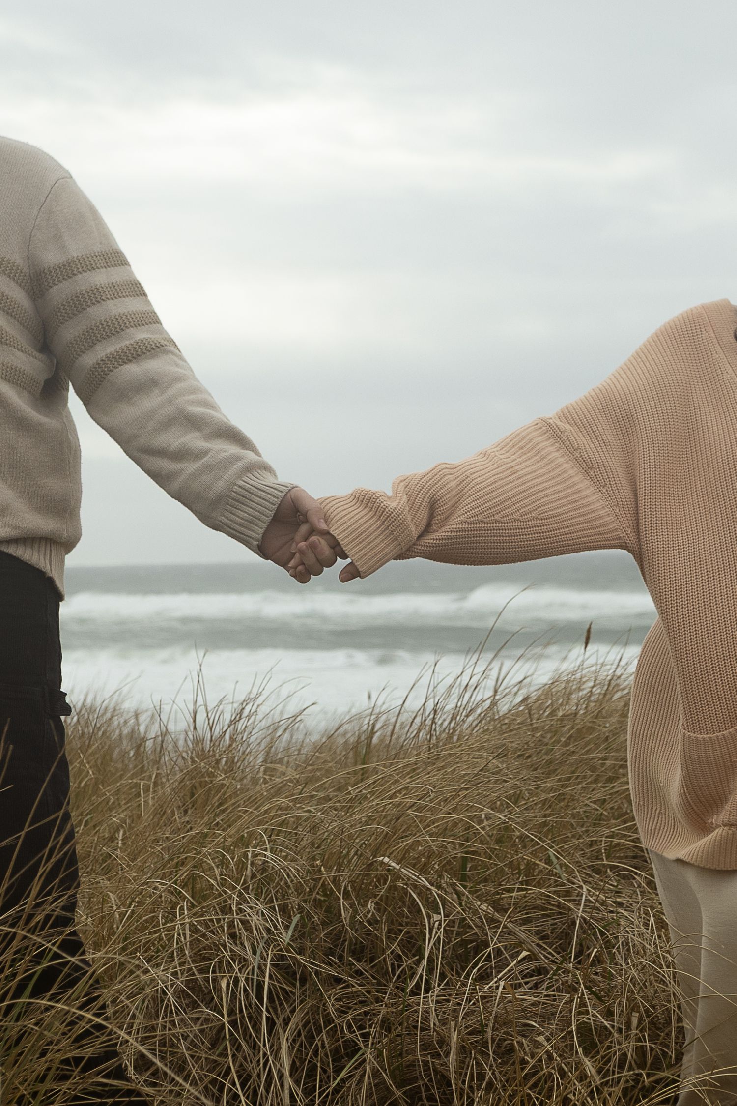 Couple holding hands on a beach with tall grass and ocean waves in the background on a cloudy day.