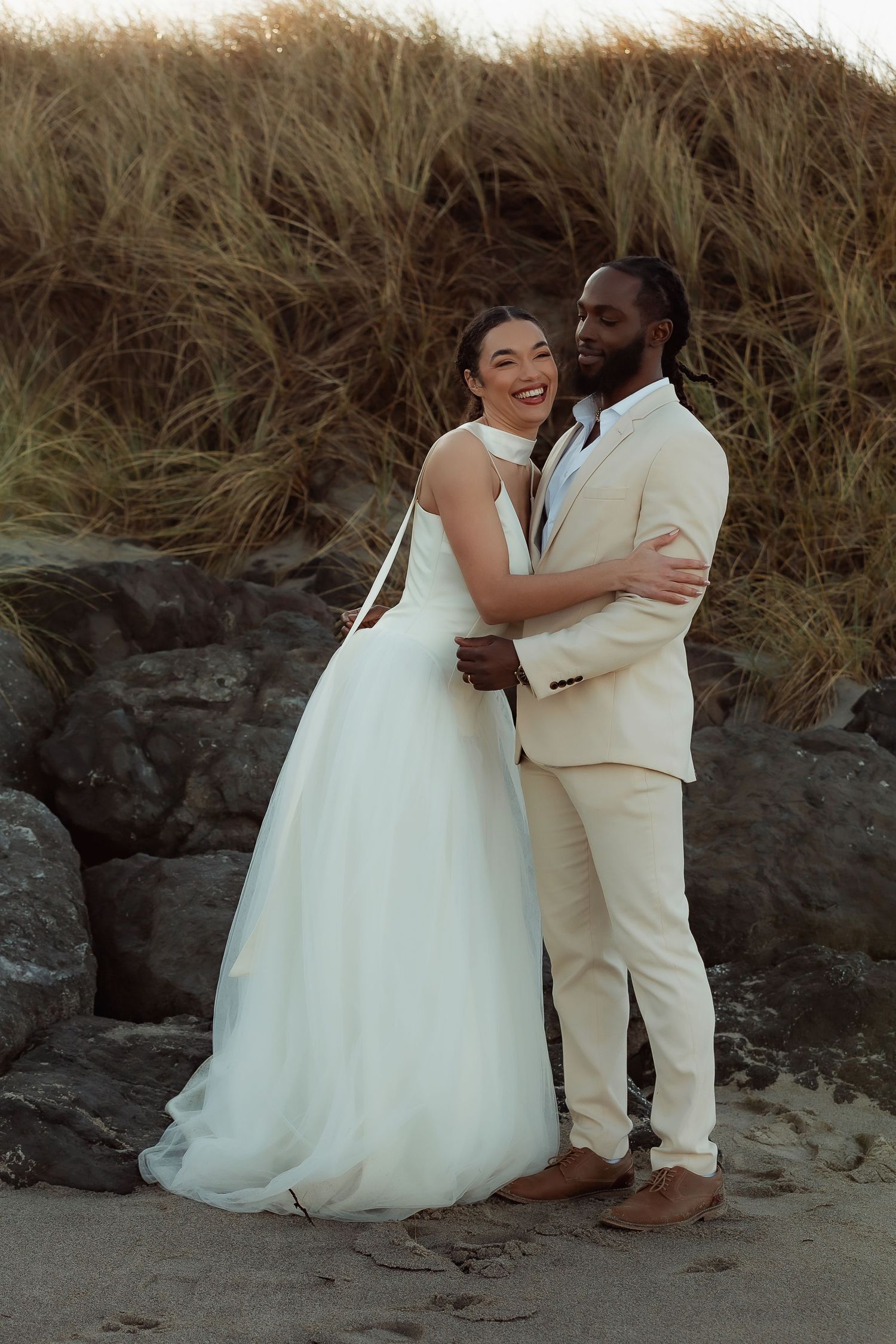 Interracial couple embracing on beach in wedding attire, bride in white gown, groom in beige suit, sandy shore backdrop.