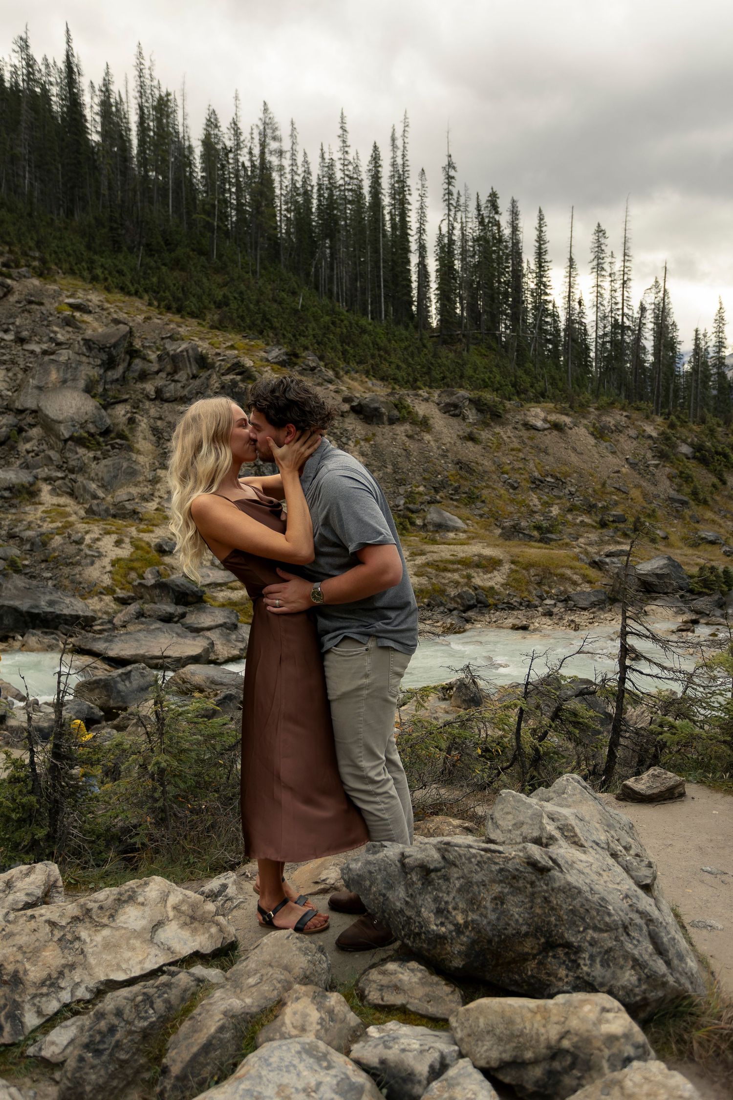 A romantic moment shared between two people on rocky terrain with evergreen trees in the background Takakkaw Falls in British Colombia.