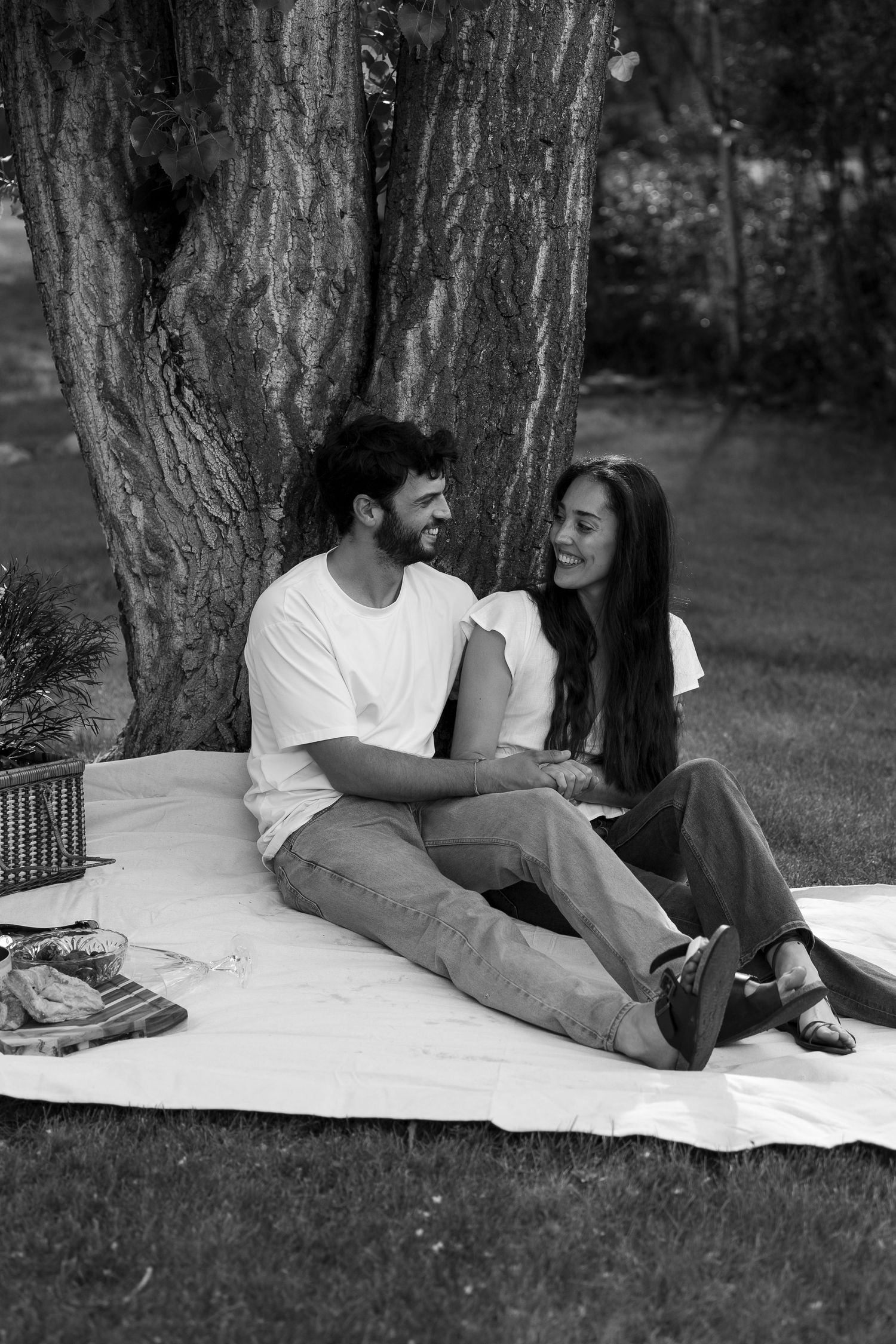 Couple relaxing on white blanket under large tree trunk in black and white photography.