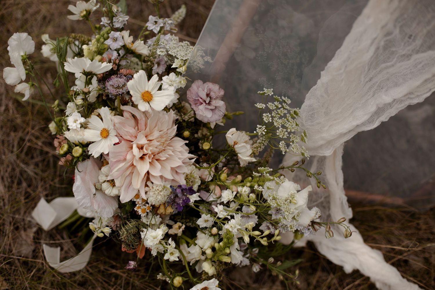 Romantic bridal bouquet with dahlias, cosmos, and wildflowers in soft pink, white, and purple hues resting on dry grass.