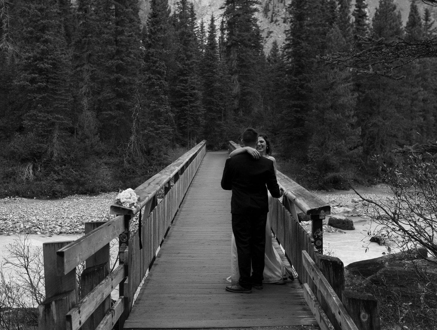 Black and white photo of a couple embracing on a wooden bridge surrounded by tall pine trees on their wedding day.