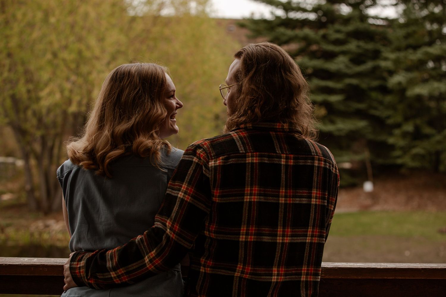 Couple sitting together on a wooden deck, sharing a tender moment surrounded by lush green trees.