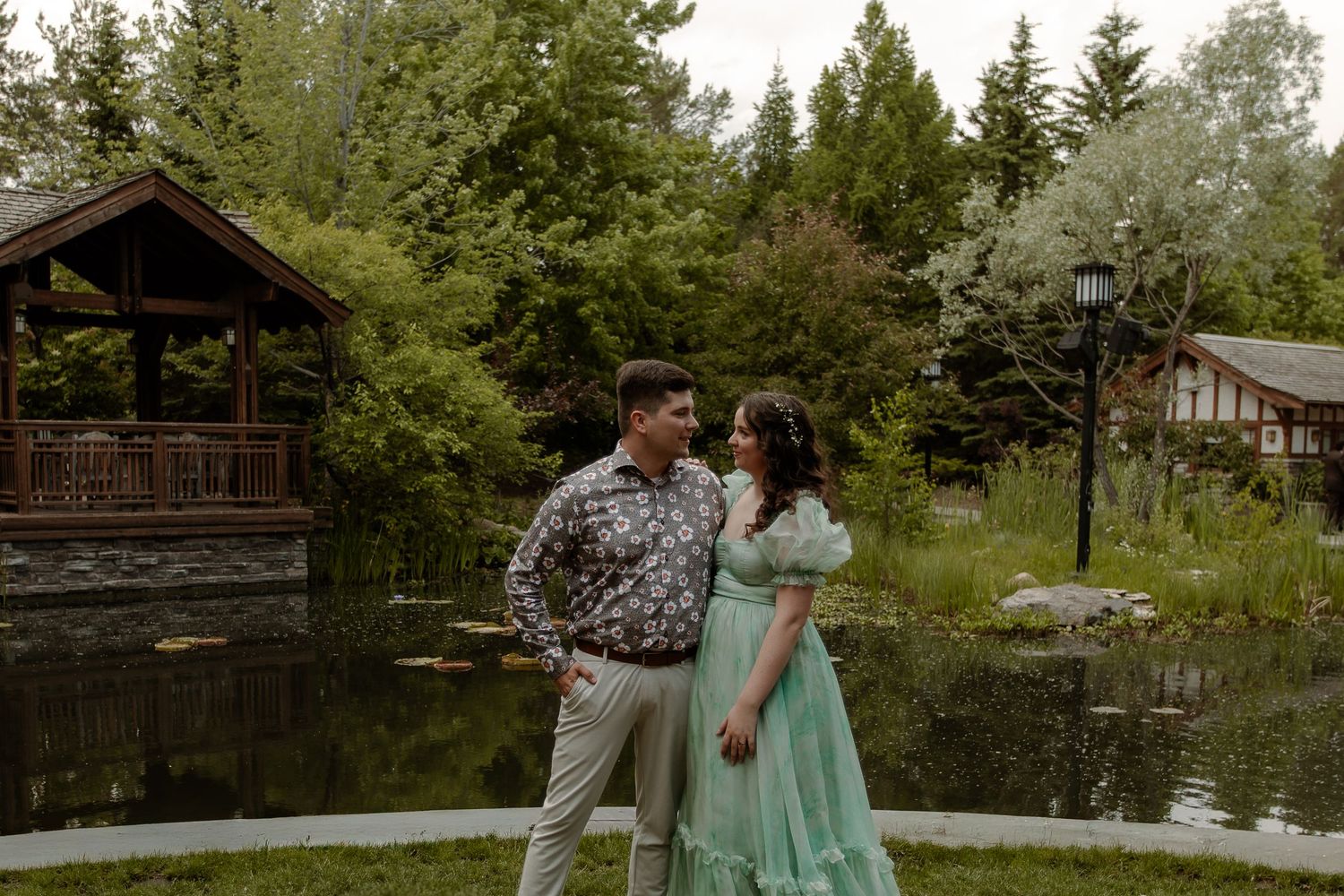 Couple in elegant attire standing by a serene pond with a wooden gazebo and lush greenery backdrop.