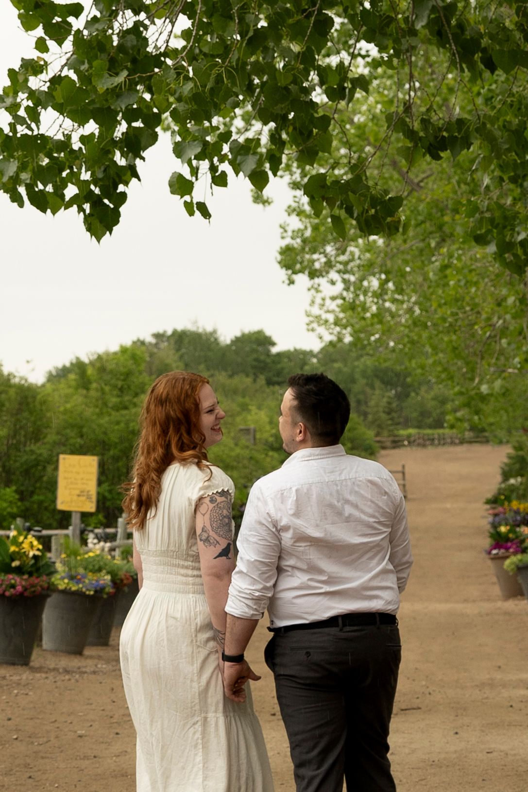 Couple holding hands walking on a garden path, woman in white dress with red hair and tattoo, man in white shirt.