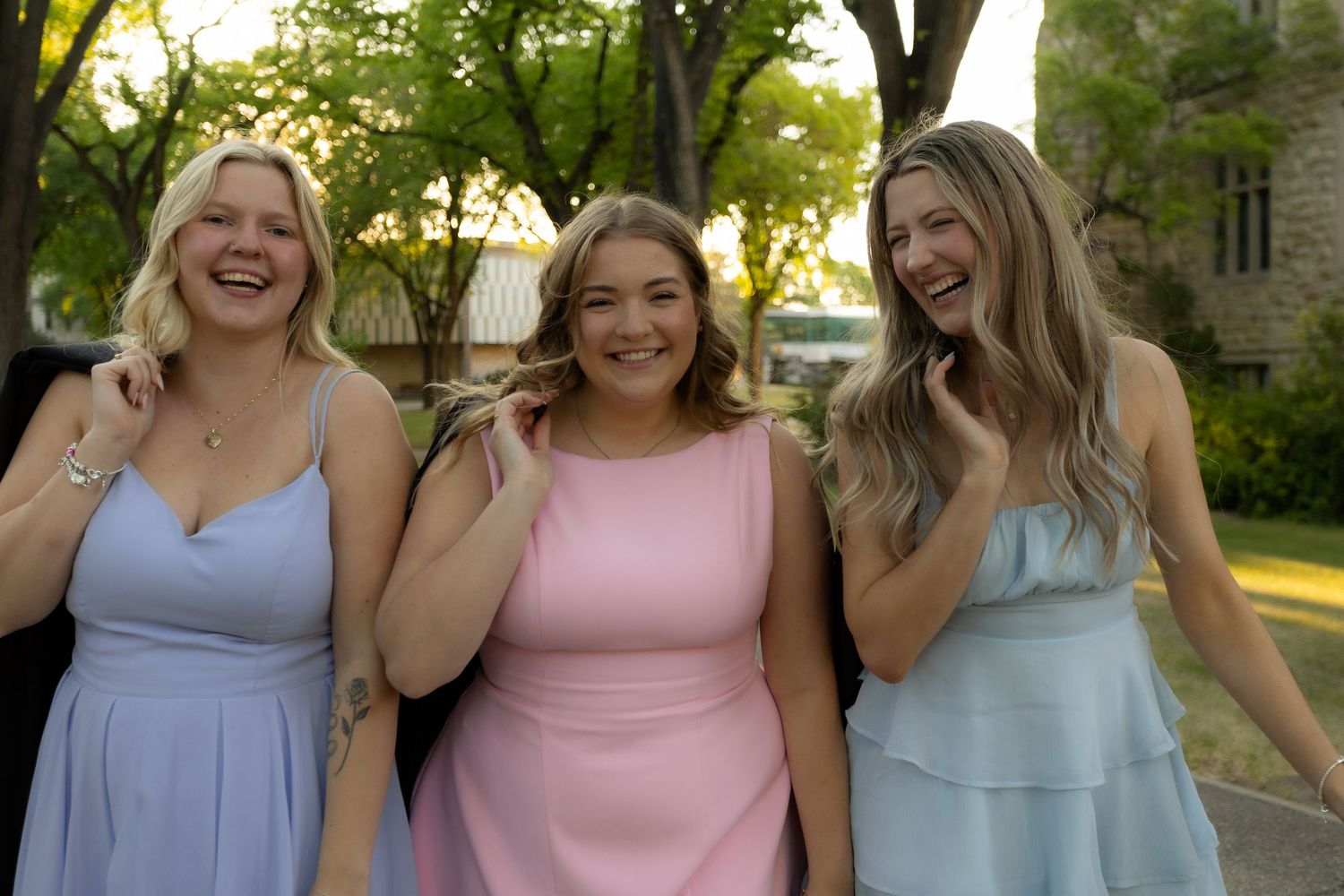 Three young women in pastel dresses laughing together outdoors at a formal event.