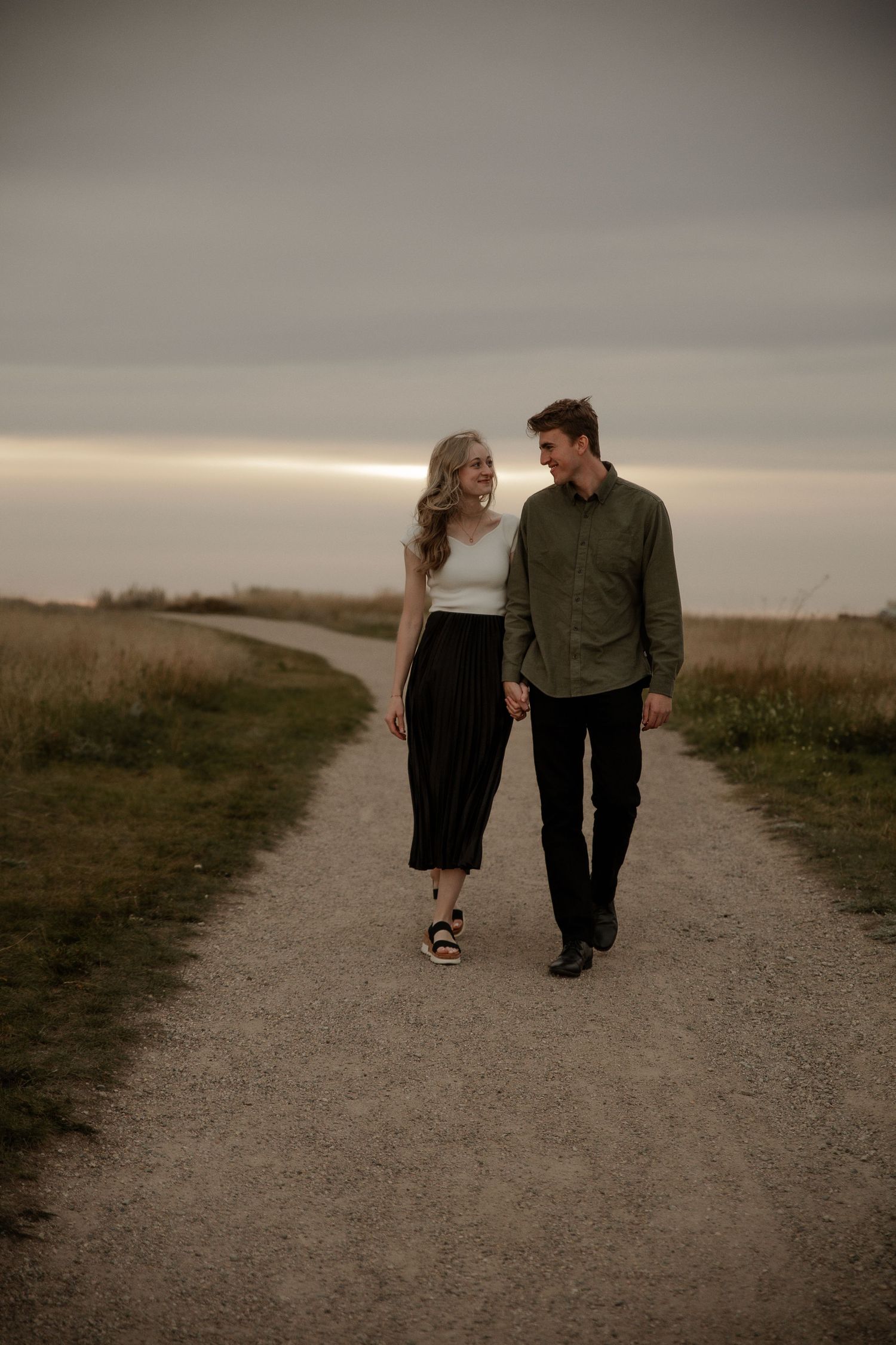 Couple holding hands walking on a gravel path through golden fields at dusk, woman in white top and black skirt.