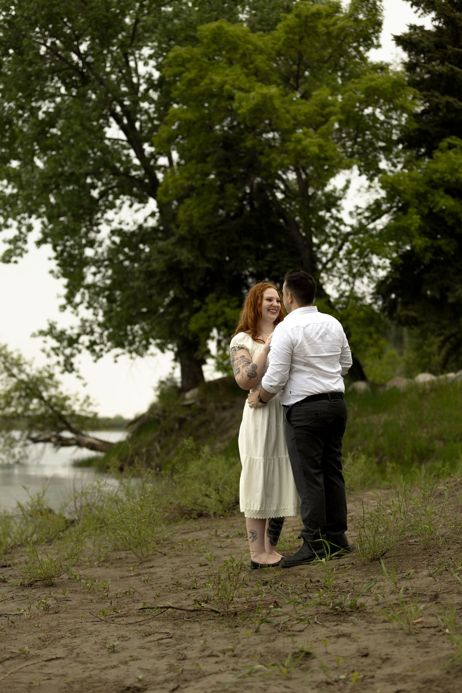 A smiling redhead woman in white dress holds hands with a man on a sandy shore near trees.