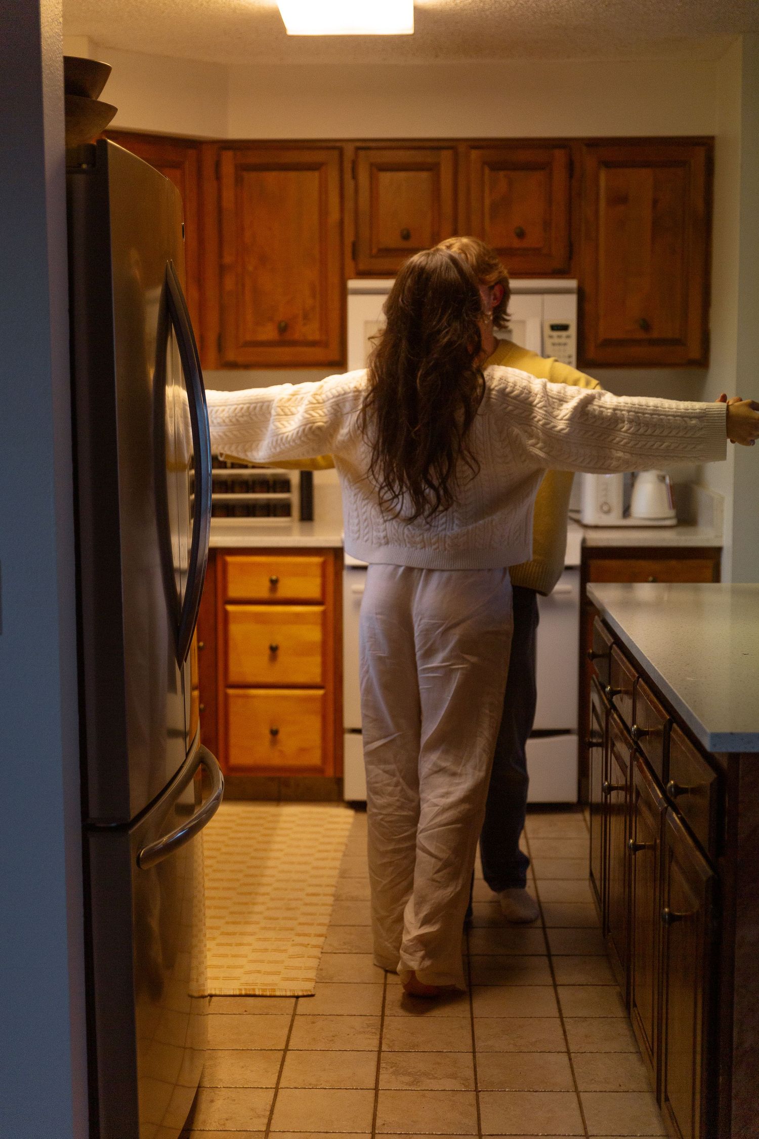 Woman in white pajamas standing in a warmly lit kitchen with wooden cabinets and stainless steel refrigerator.