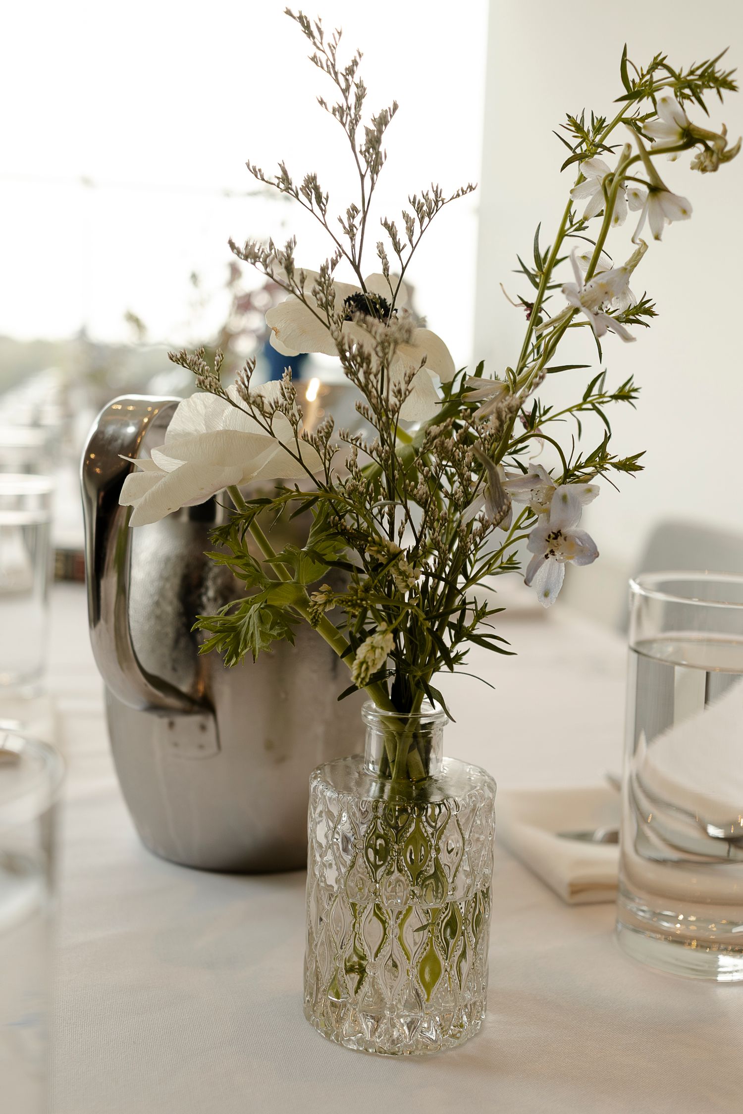 Crystal vase with white wildflowers and greenery on a white table beside a silver pitcher.