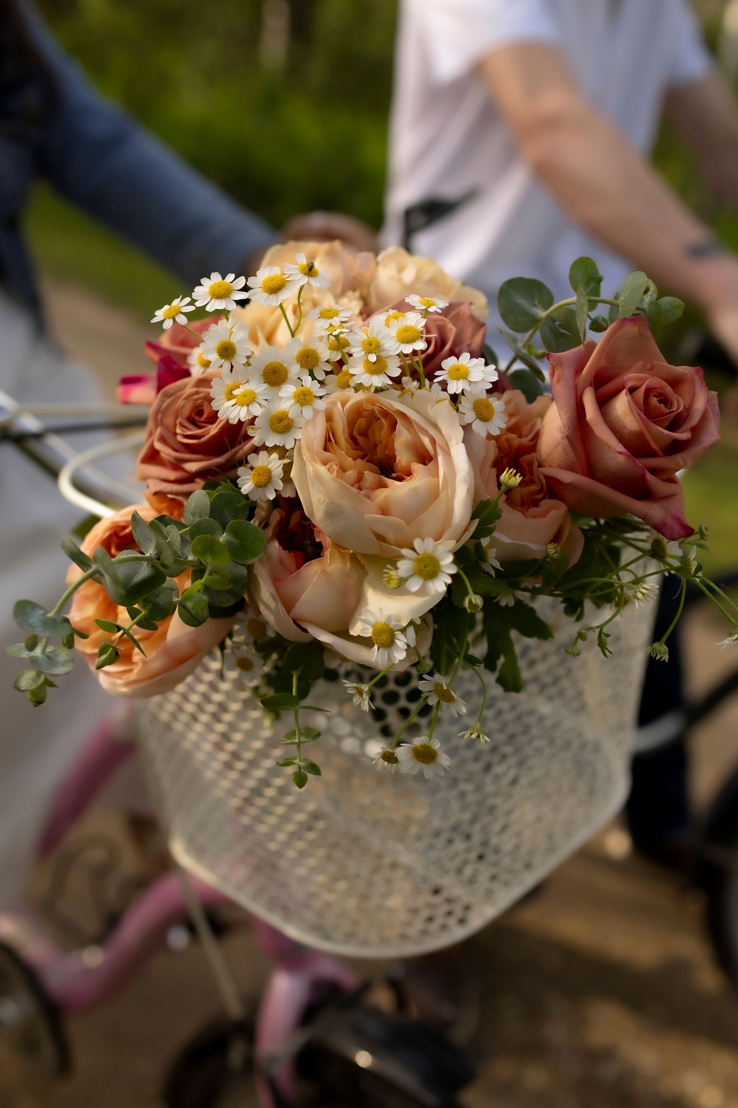 White wicker bicycle basket filled with peach roses, daisies and greenery against a blurred background.
