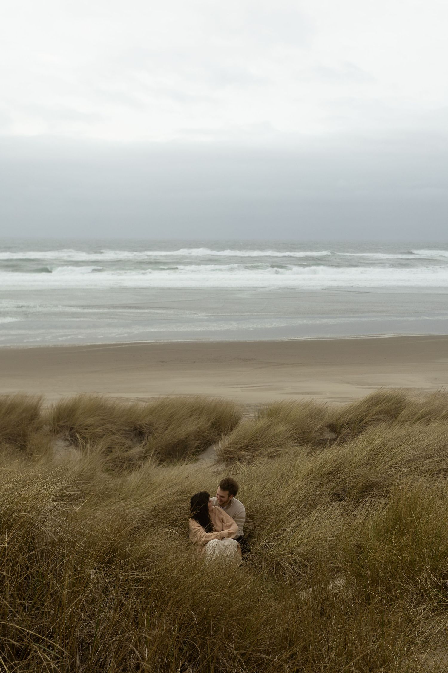 Couple embracing in coastal dune grass with stormy ocean waves and overcast sky in the background.