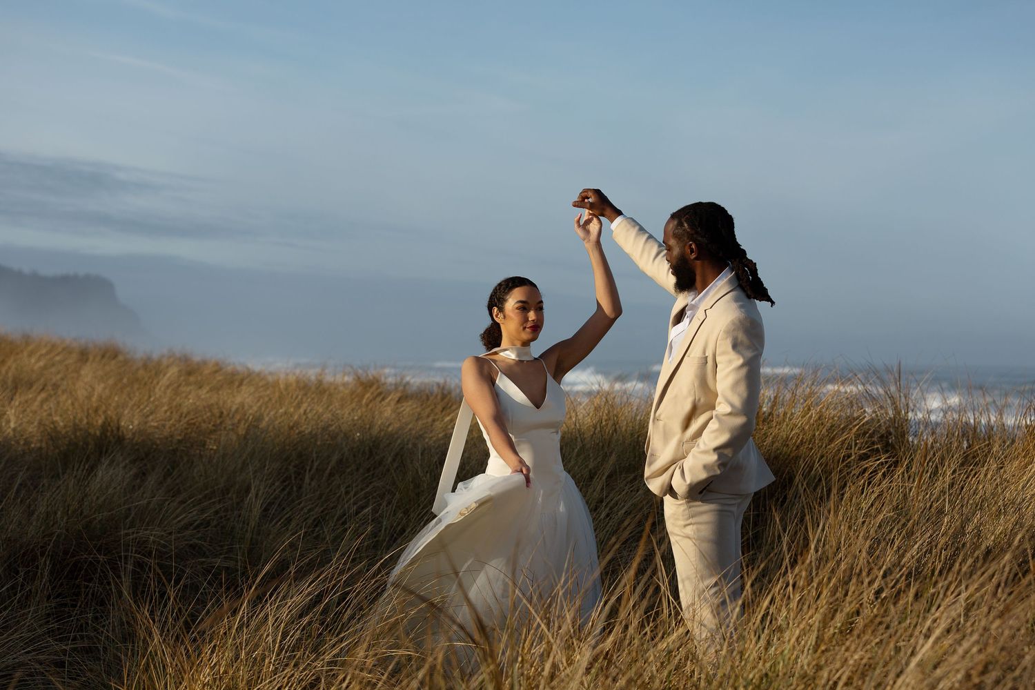 Couple dancing in golden beach grass during wedding shoot with ocean backdrop on a clear day.