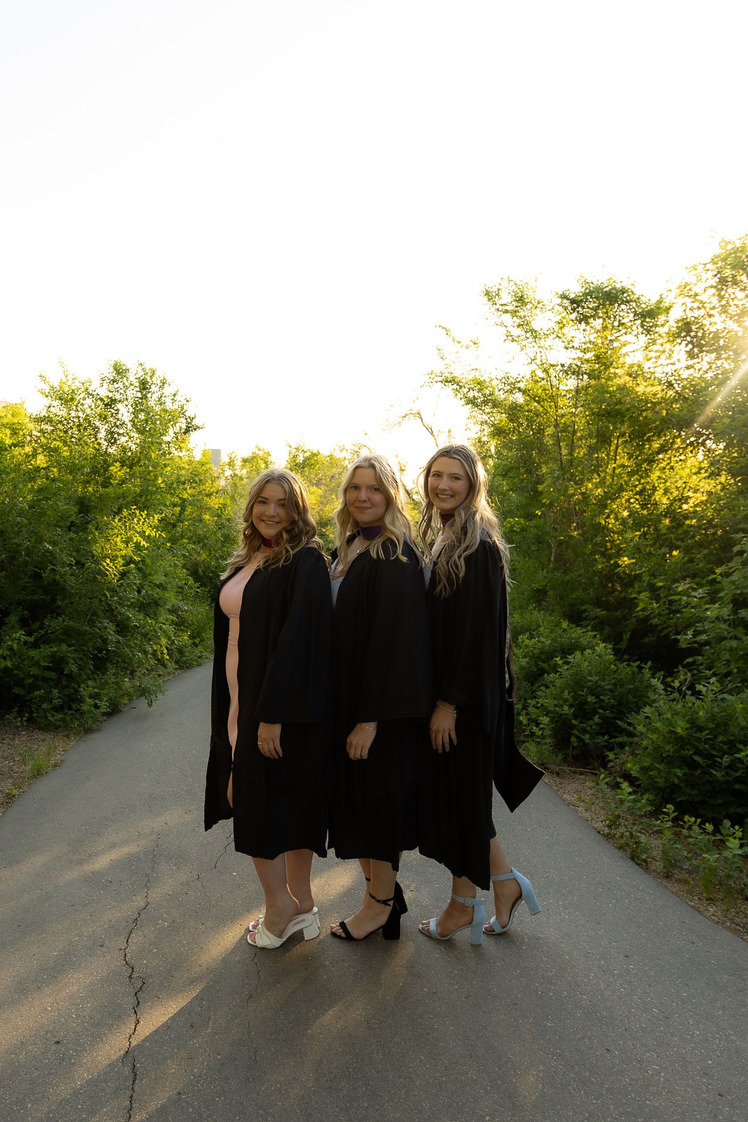 Three women in black graduation gowns and heels pose on a tree-lined path in golden hour sunlight.