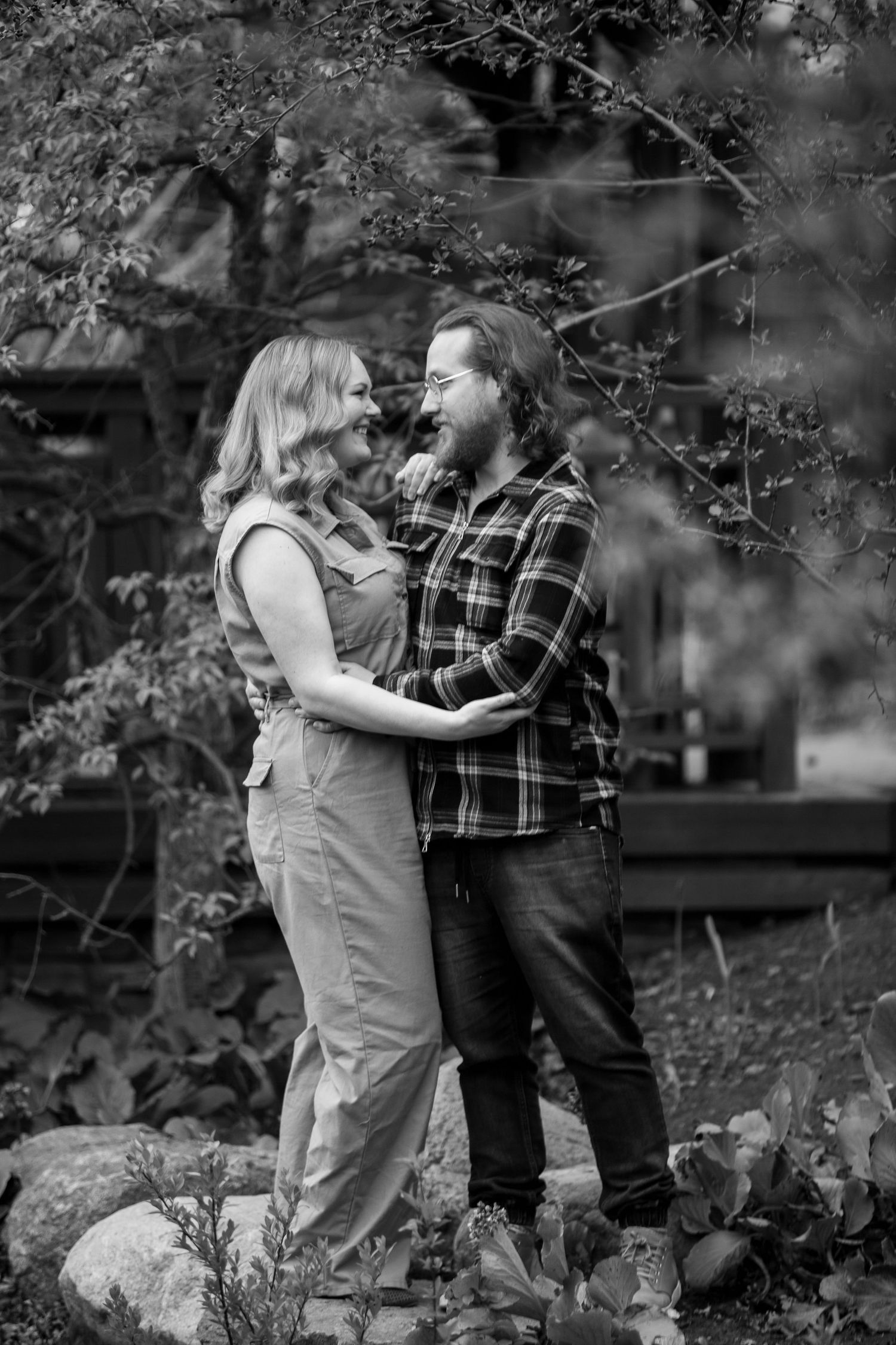 Black and white photo of a laughing couple embracing outdoors among lush trees and rocks.