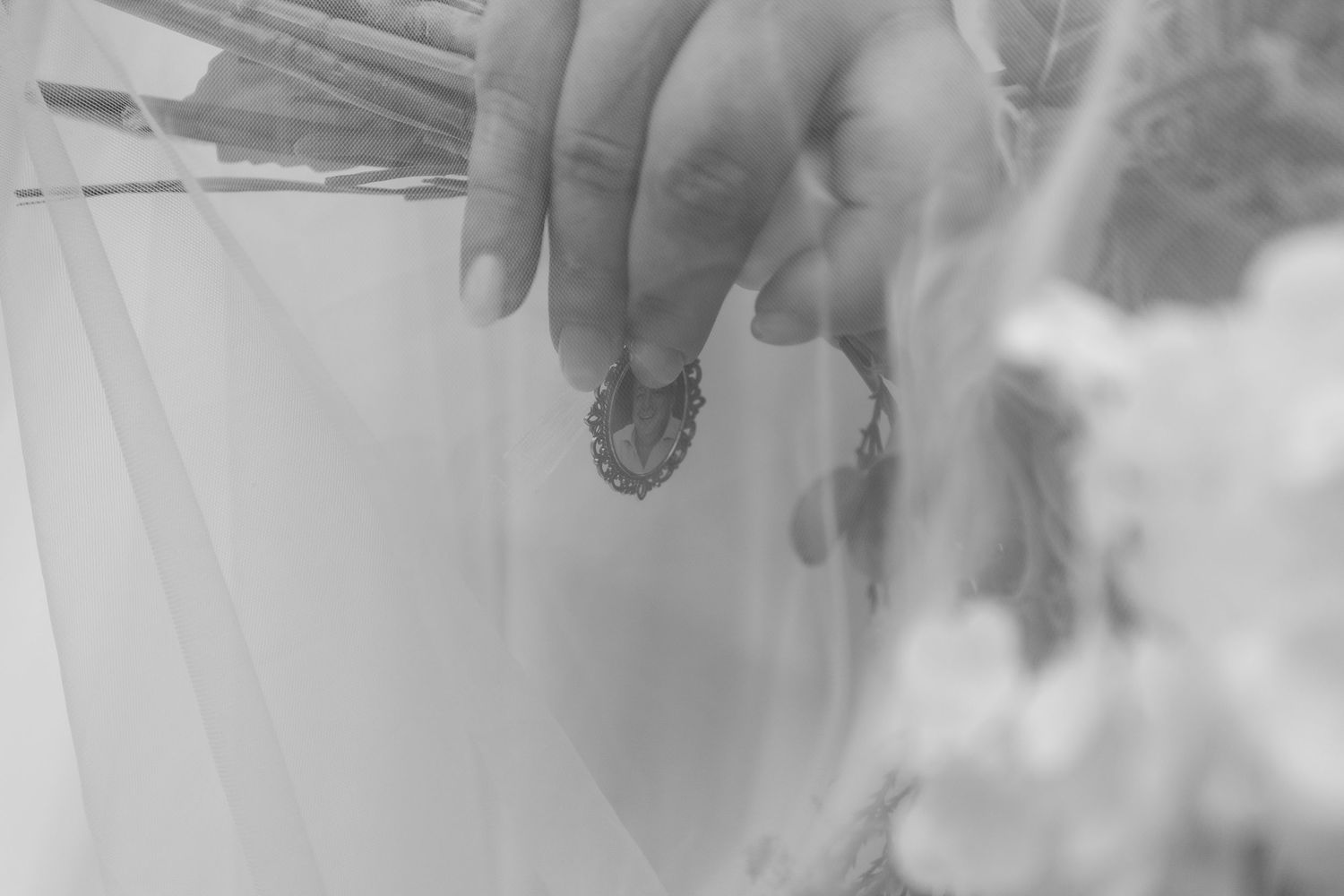 Black and white photo of a hand holding a delicate heart-shaped pendant near a floral bouquet.
