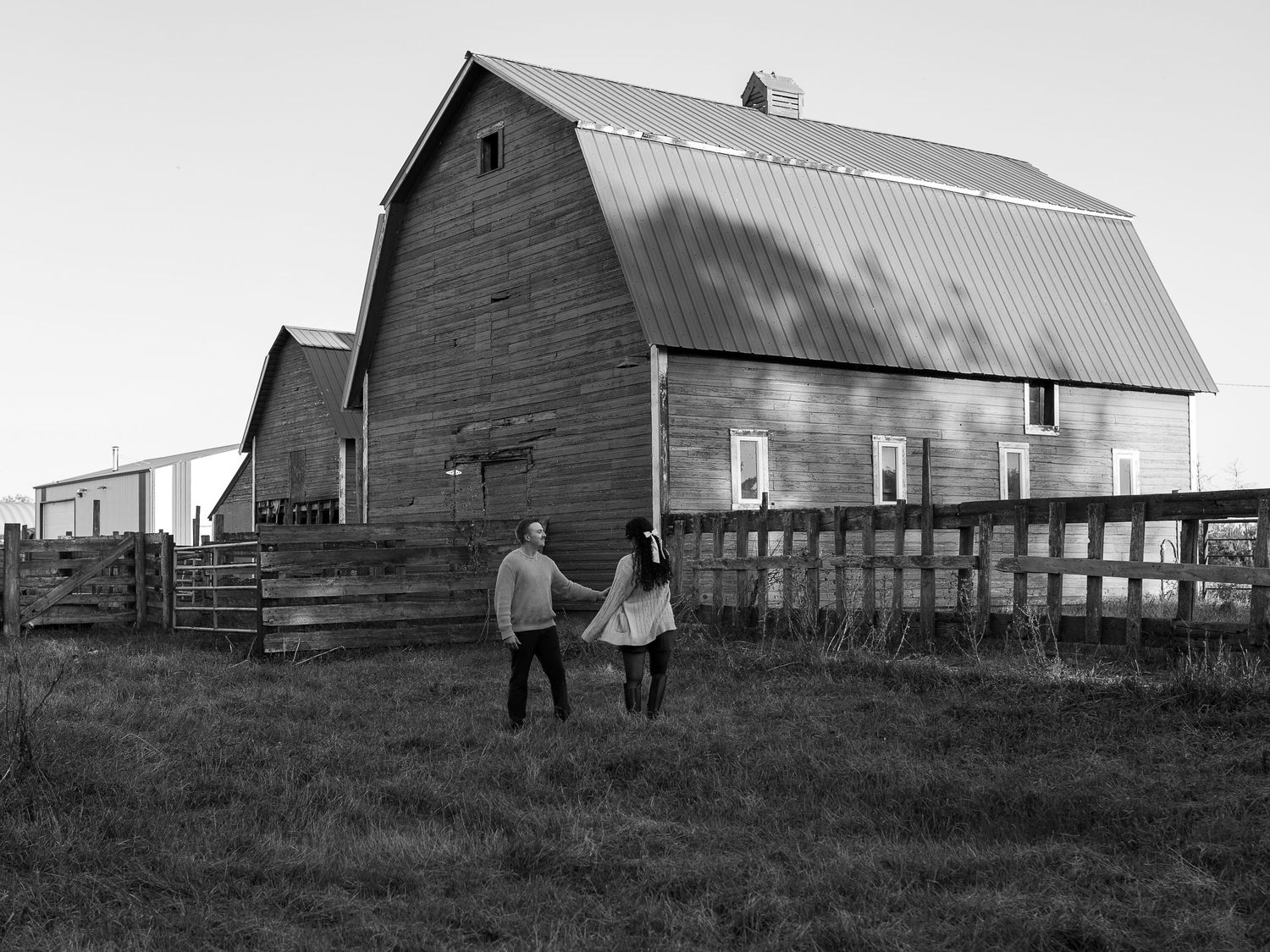 A couple walking hand in hand towards a rustic barn in black and white photography on a farm in the Canadian prairies.
