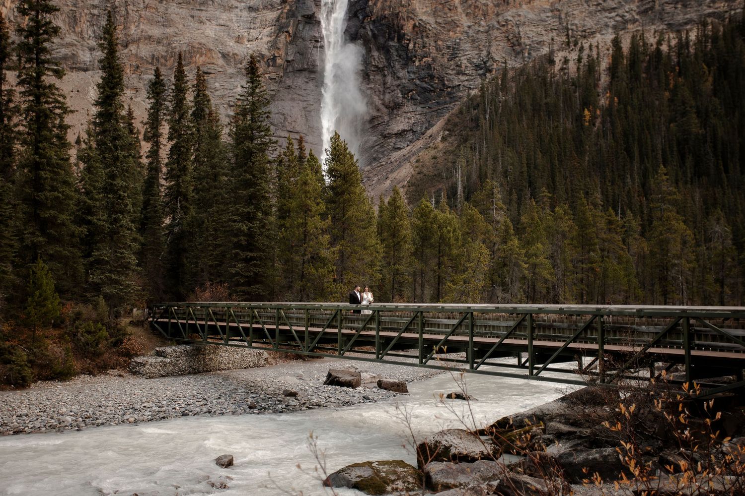 A couple stands on a wooden bridge with a dramatic waterfall cascading in the background at Banff National Park.