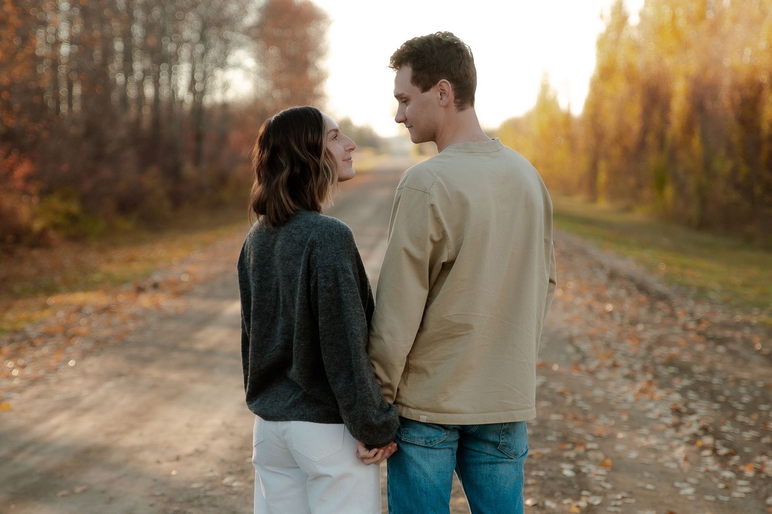 Couple holding hands on a scenic autumn dirt road, surrounded by golden trees in warm sunset light.