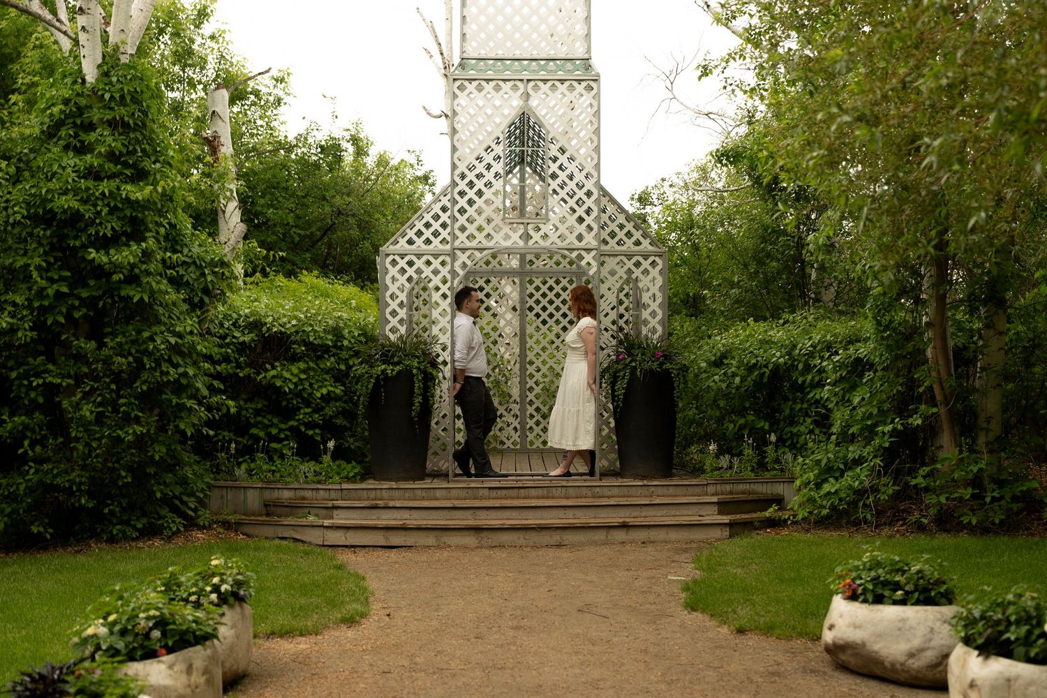 Couple standing before a white lattice garden chapel surrounded by lush green trees on a cloudy day.