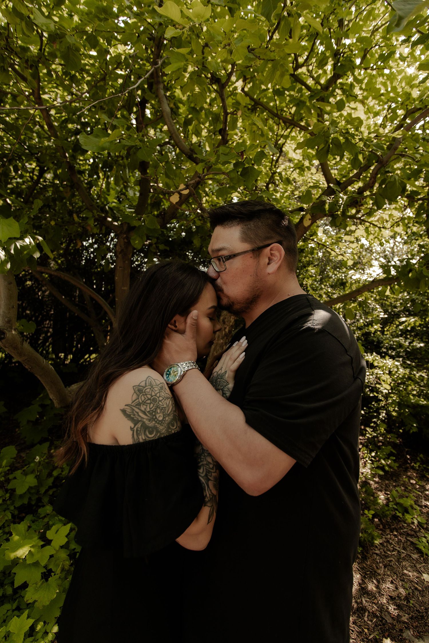 A couple shares a tender kiss surrounded by lush green trees, both wearing black outfits with visible tattoos.