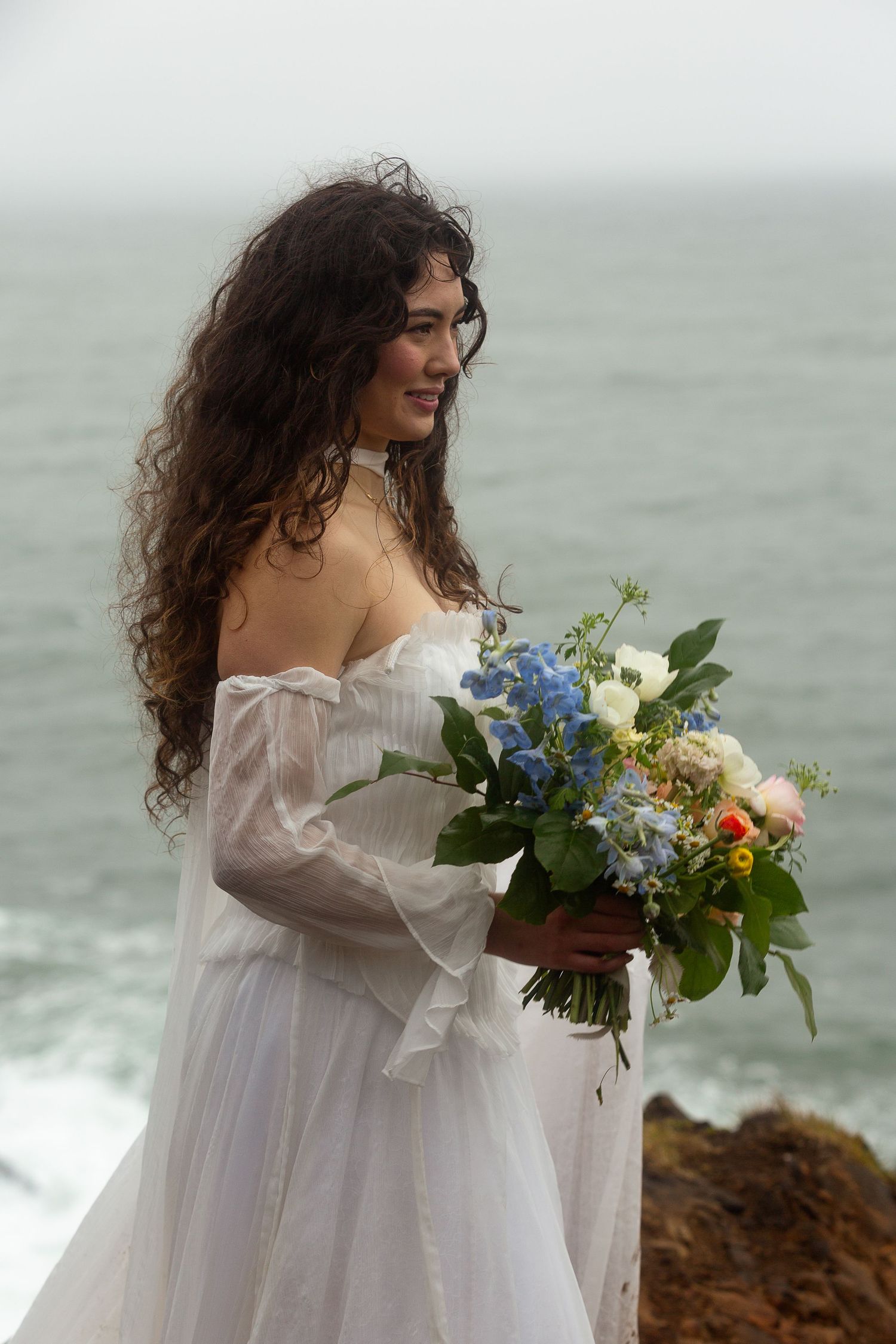 Bride in white off-shoulder gown holds blue and white bouquet on rocky ocean cliff.