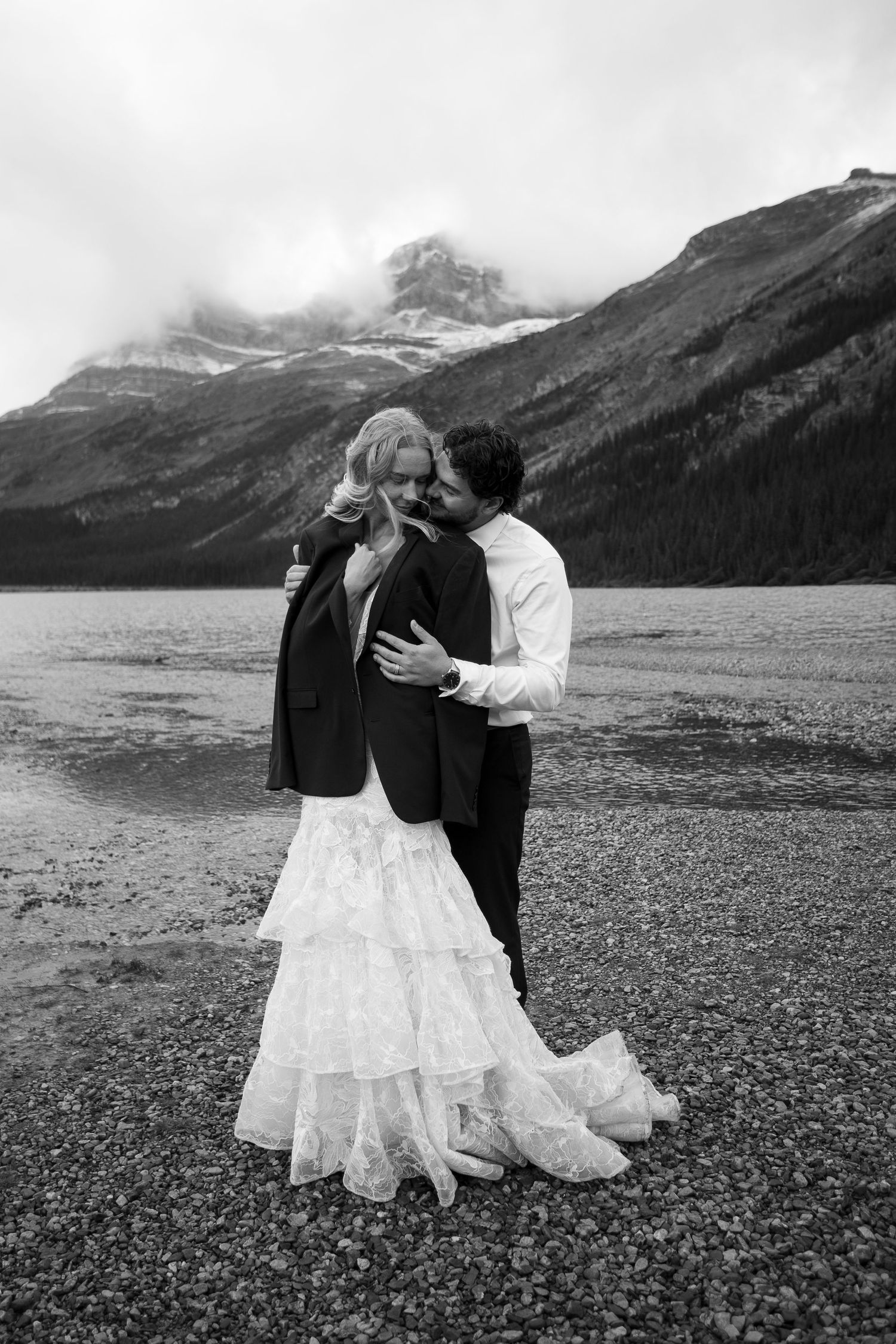 Black and white photo of a bride and groom embracing by a mountain lake, with snow-capped peaks in the background.