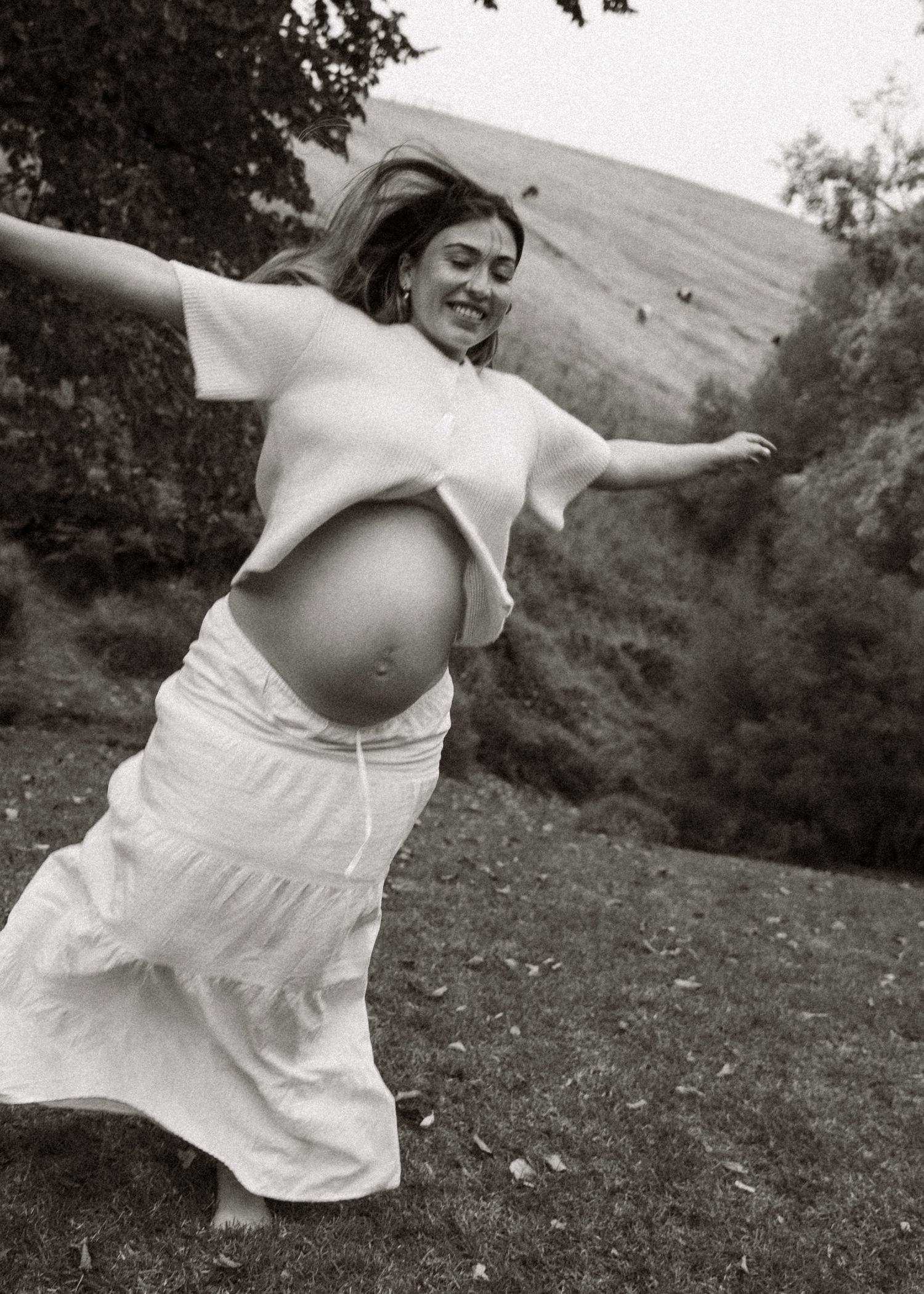 A pregnant woman dancing in a field at the end of her pregnancy in the hills at sunset at a maternity family photography session.
