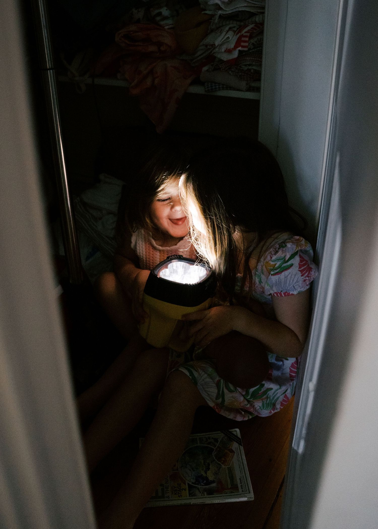 Young girls laughing in a closet using a torch under their chin playing pretend during a photography session at home.