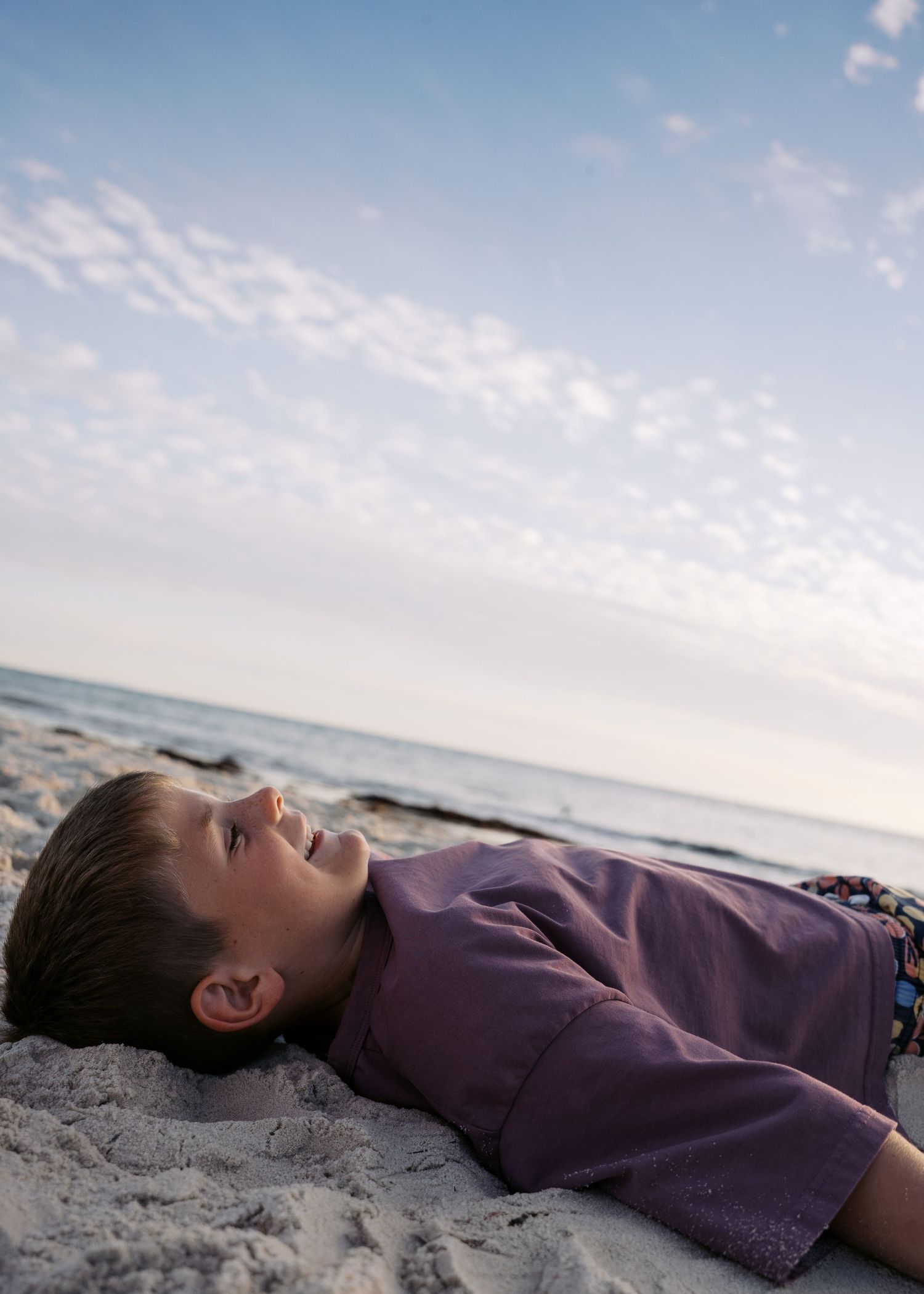 A young boy laying on the sand at the beach, looking up at the clouds at sunset during a family photography session.