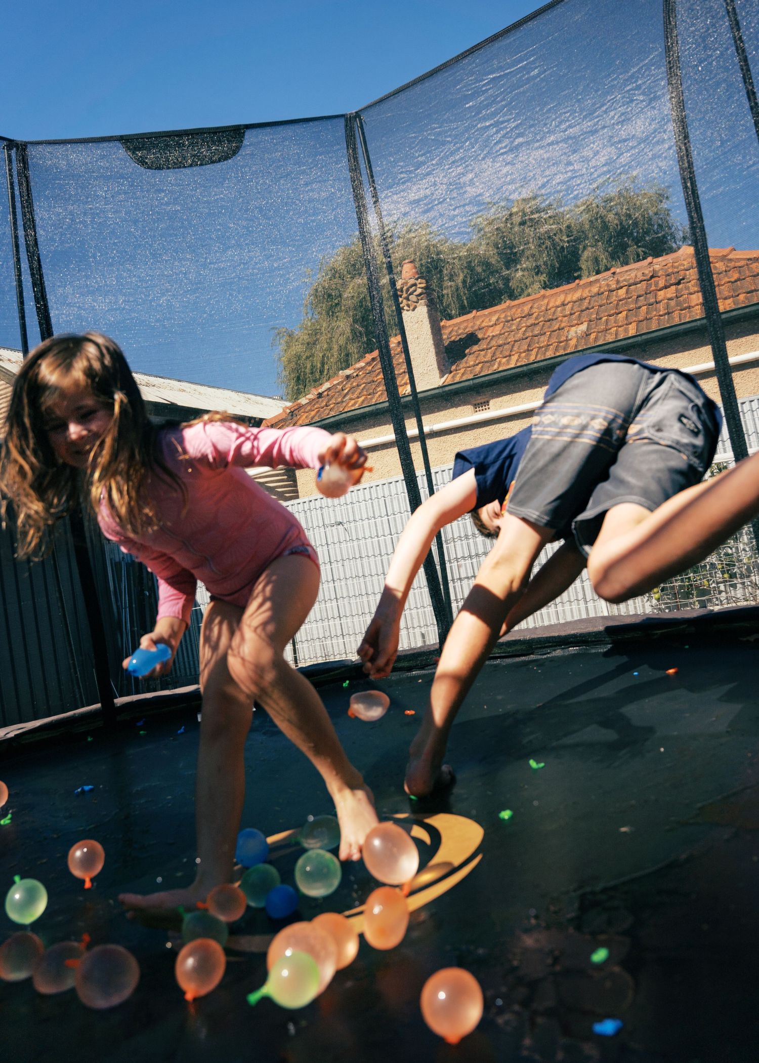 Siblings having a water fight with water balloons on a trampoline during a family photoshoot at home.