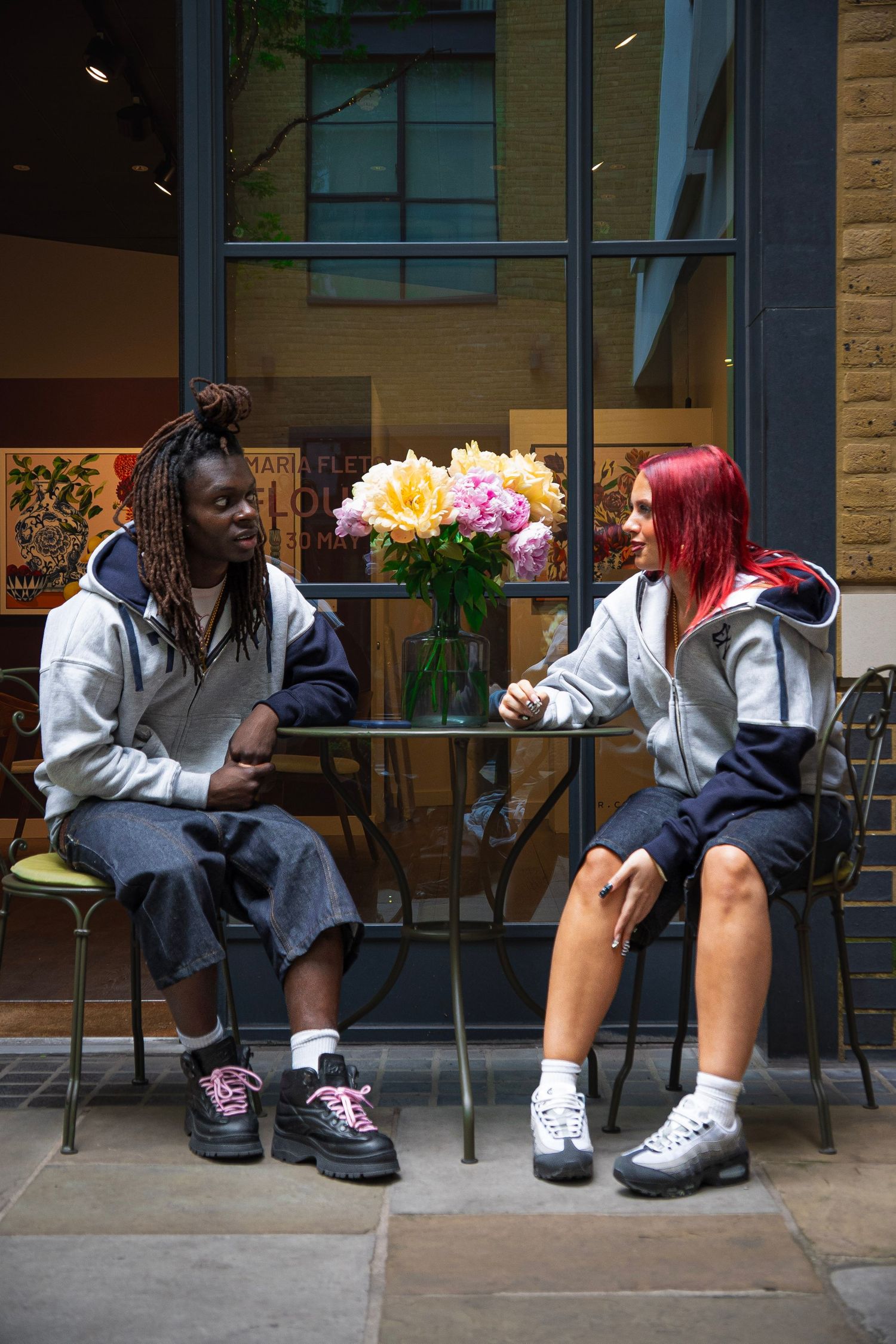 Two friends in matching gray hoodies sit at an outdoor cafe table with pink flowers, wearing chunky sneakers.