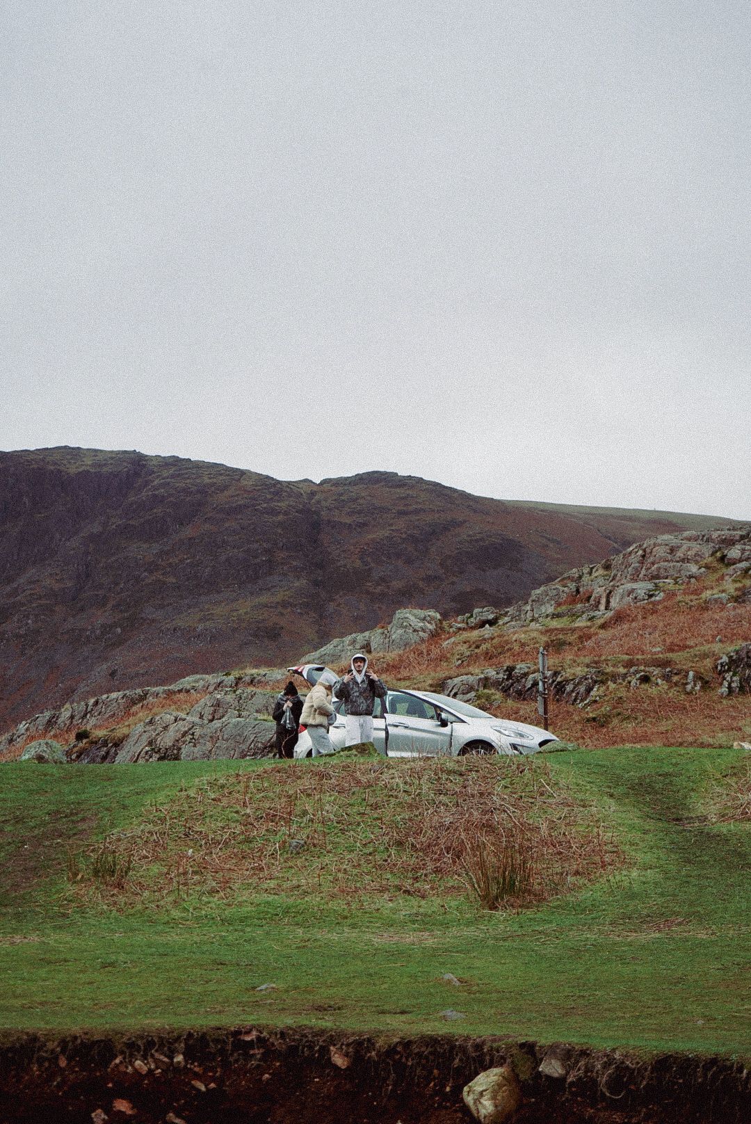 Two people stand beside a silver car parked on a grassy hill with rocky mountains behind.