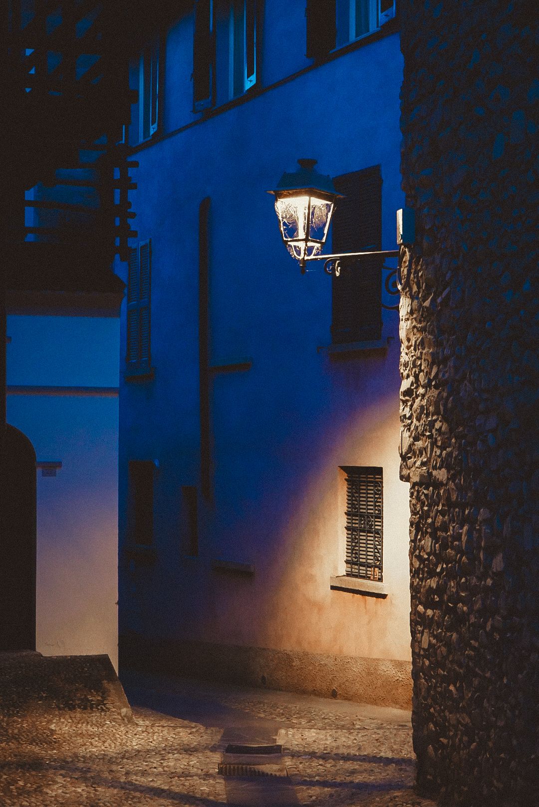 Glowing vintage street lantern illuminates cobblestone alley between stone and plastered walls at night.